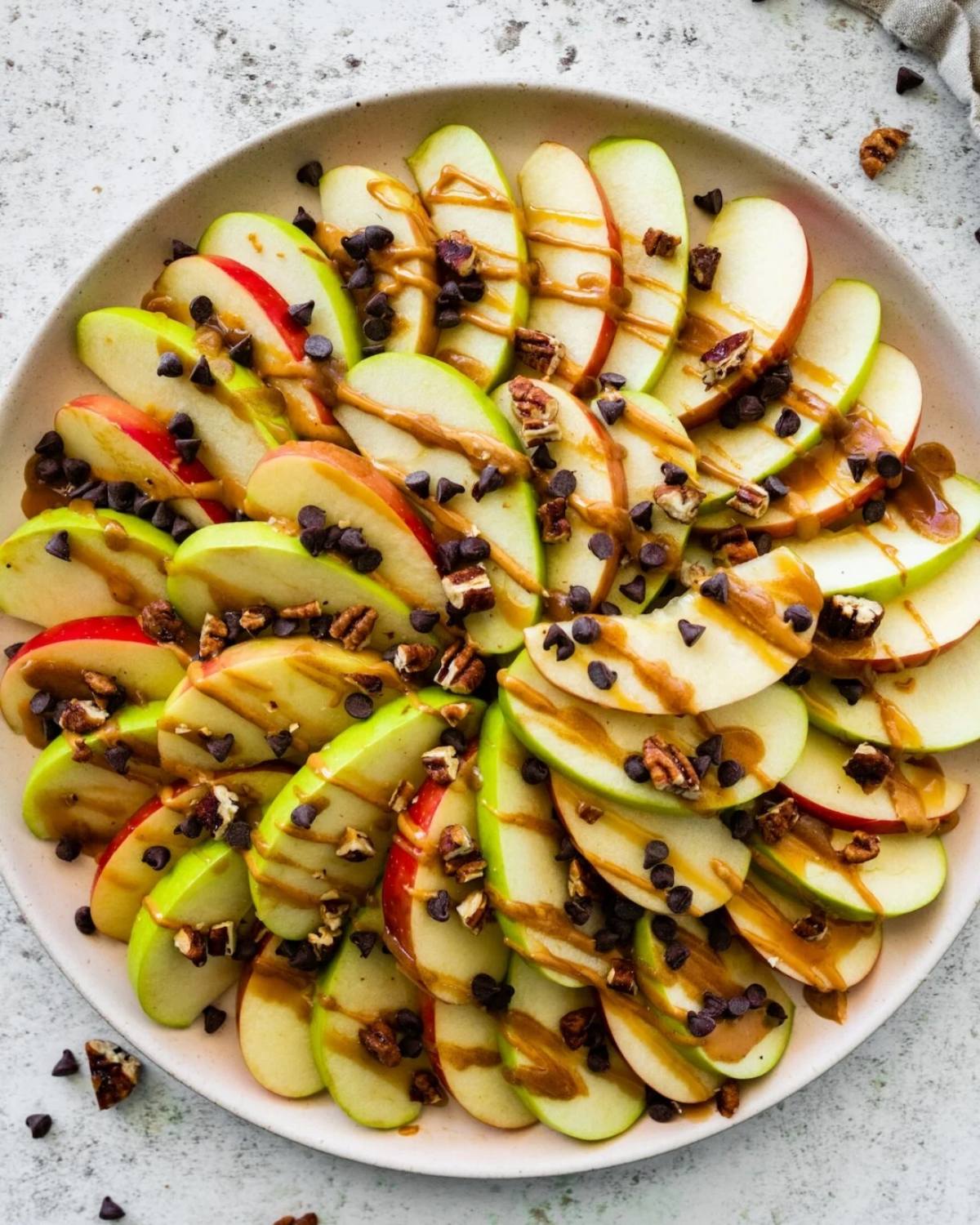 Healthy fall dessert apple nachos arranged on a plate with sliced red and green apples, peanut butter drizzles, chocolate chips, and chopped pecans