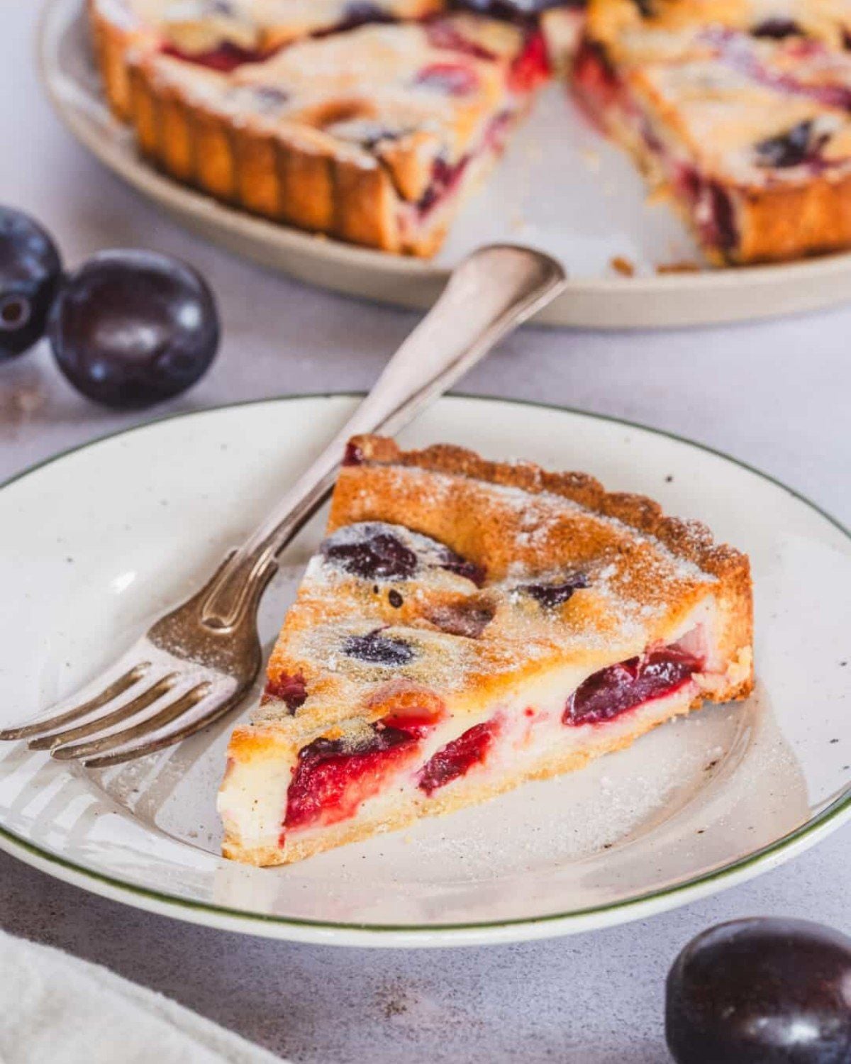 Healthy fall dessert slice of plum tart with a creamy filling and fresh plums on a white plate with a fork