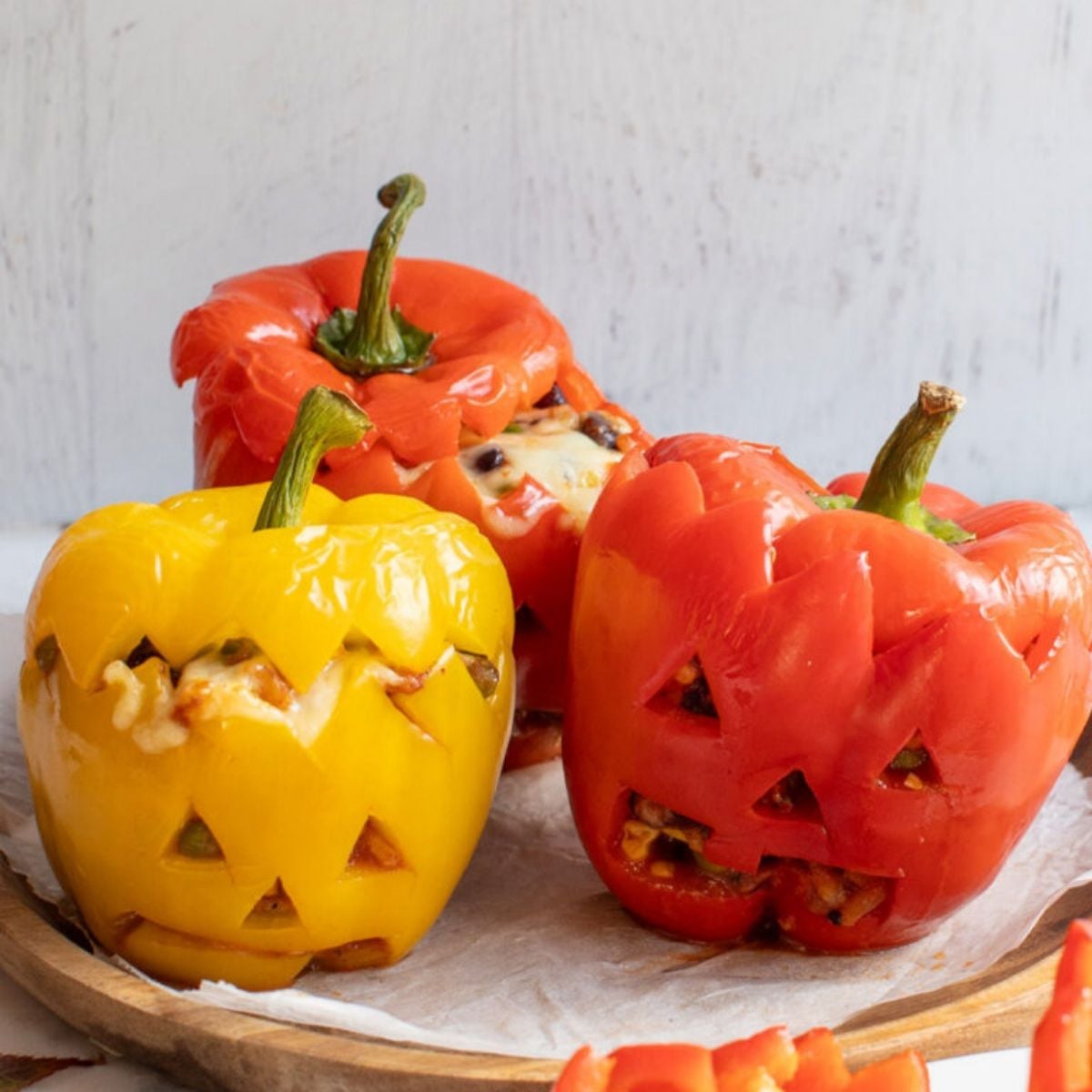 Healthy Halloween treats featuring colorful red and yellow bell peppers carved into jack o' lantern faces and stuffed with Mexican-style ingredients on a wooden tray.