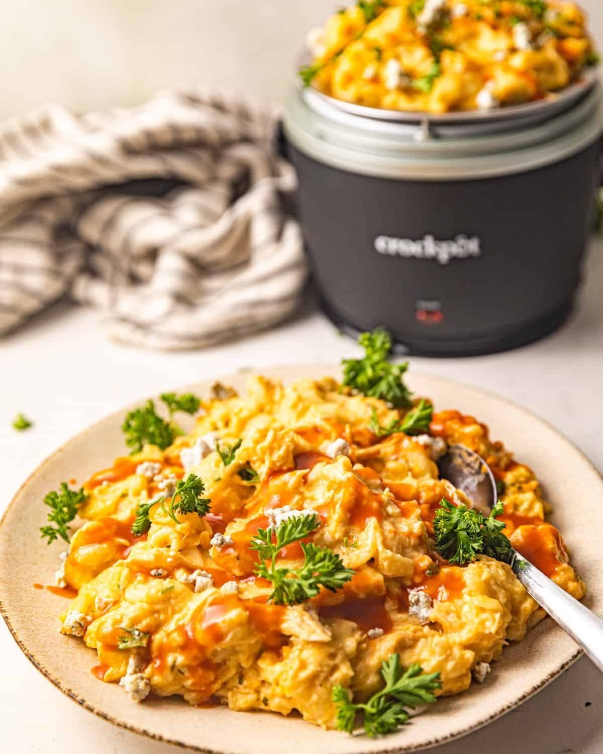 Slow cooker high-protein buffalo chicken mac and cheese on a plate topped with buffalo sauce, blue cheese crumbles, and parsley, with a crockpot in the background.