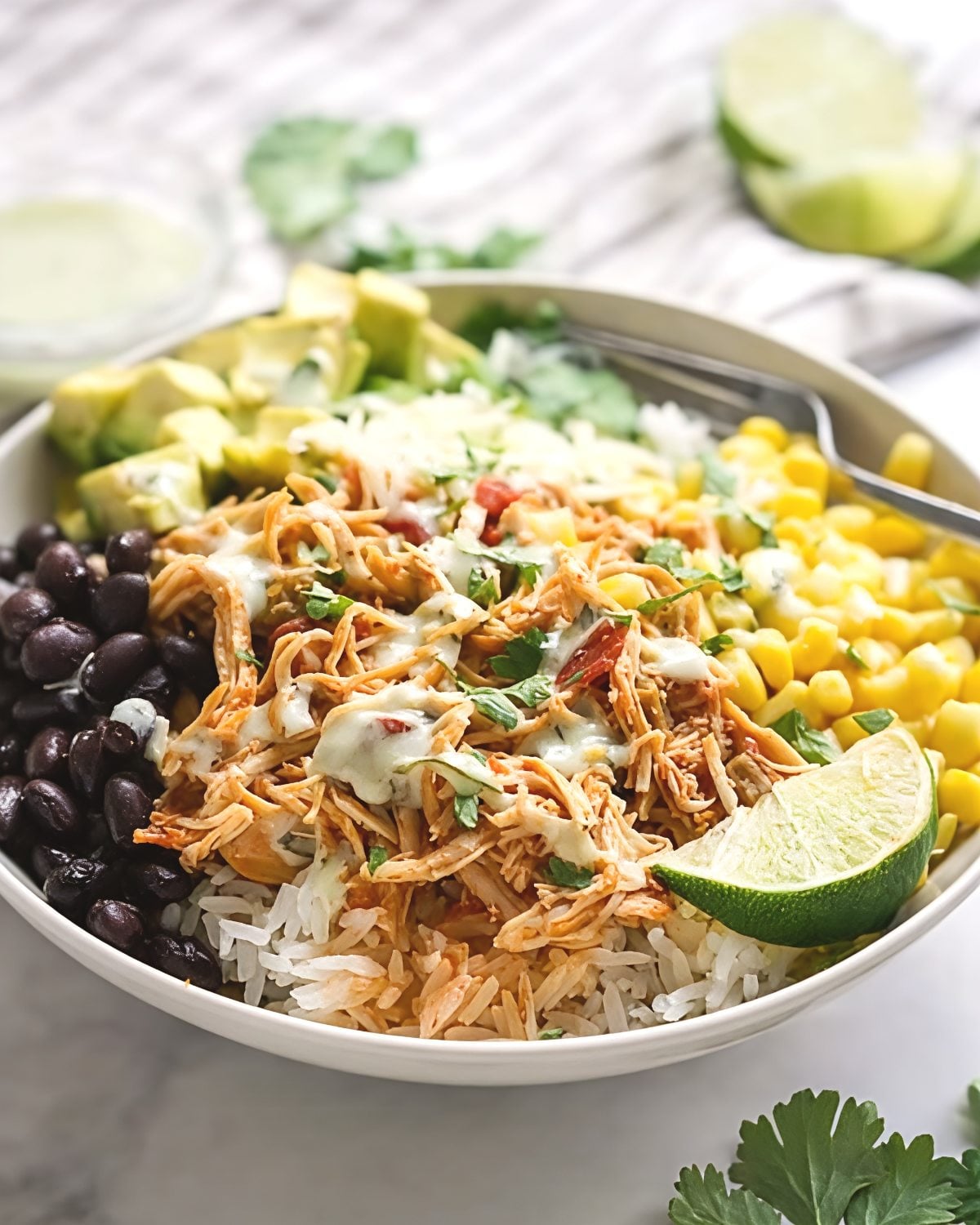 A white bowl filled with shredded slow-cooker chicken over white rice, surrounded by black beans, sweet corn, cubed avocado, shredded cheese, and a lime wedge, drizzled with creamy sauce.
