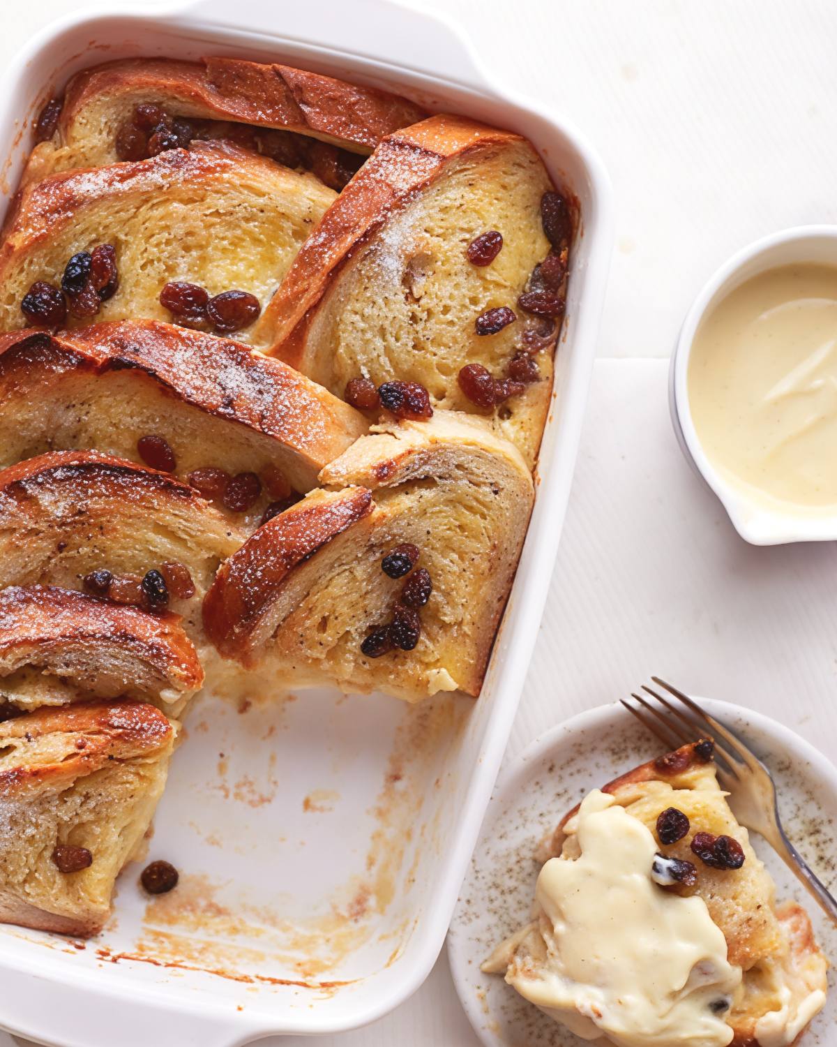 A white baking dish filled with golden bread and butter pudding studded with raisins and dusted with powdered sugar, served alongside a small plate drizzled with vanilla sauce