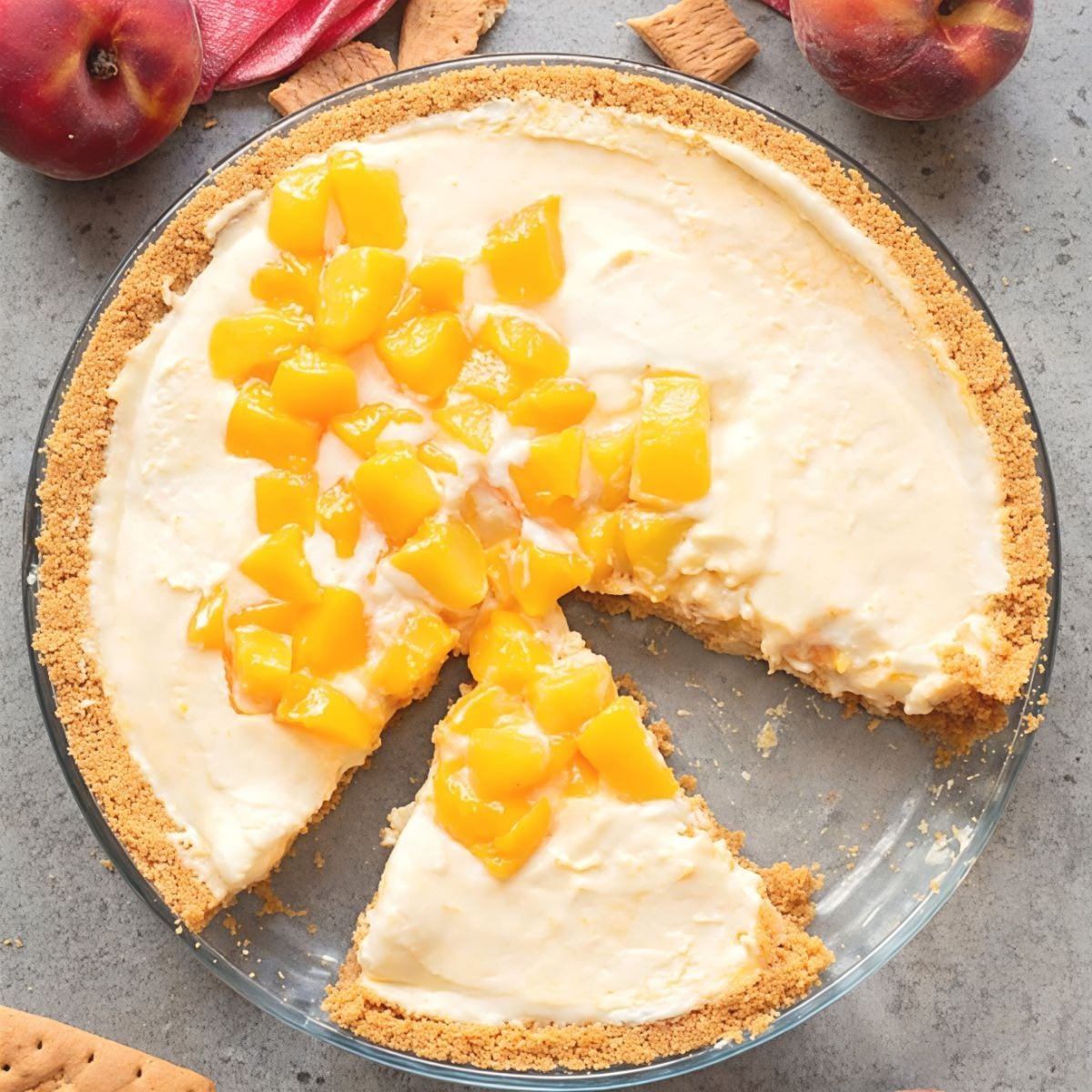 Top-down view of a peach icebox pie in a graham cracker crust with a slice pulled forward and whole peaches nearby