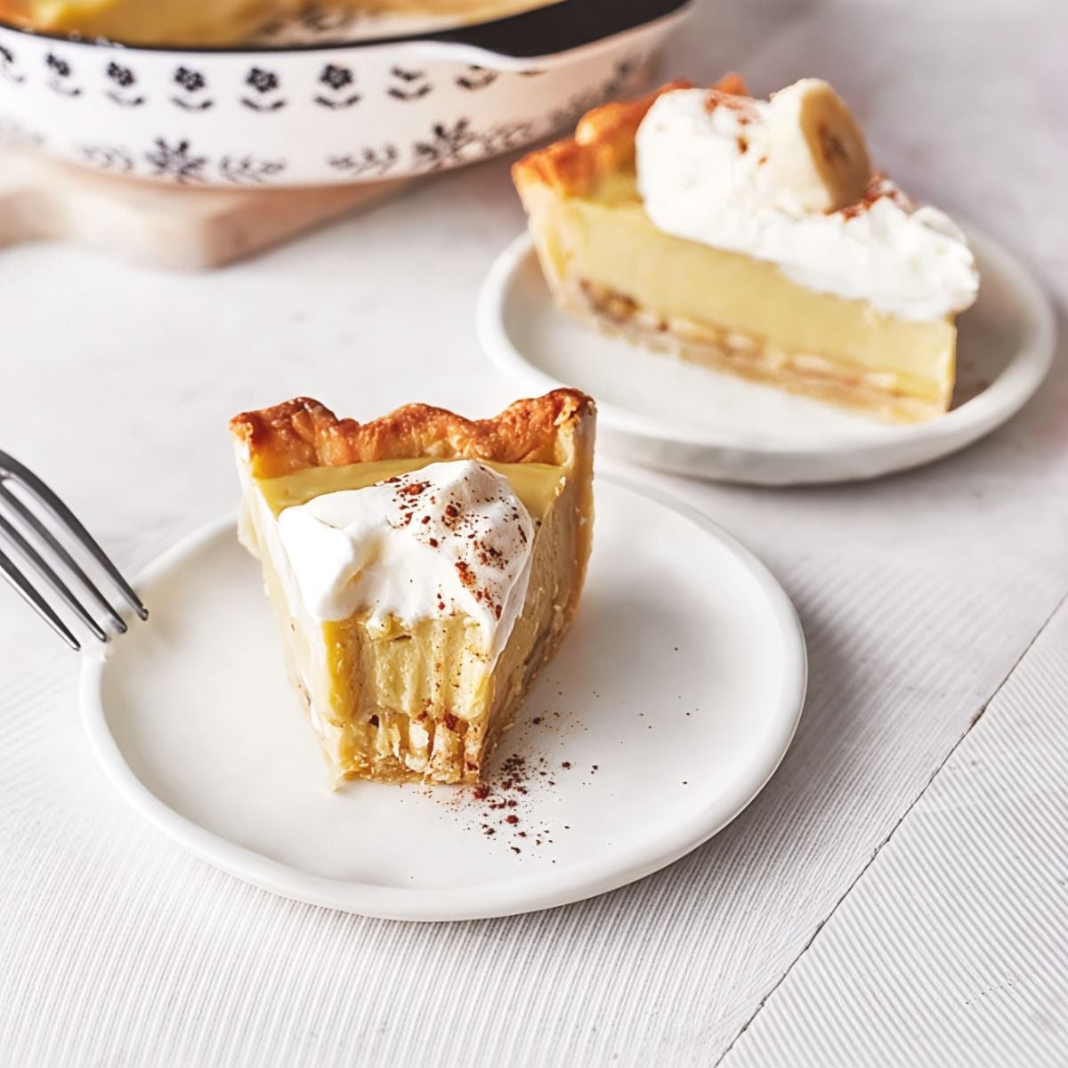 Two slices of banana cream pie in golden pastry crusts are served on white plates, topped with dollops of whipped cream, cinnamon dusting, and fresh banana slices.
