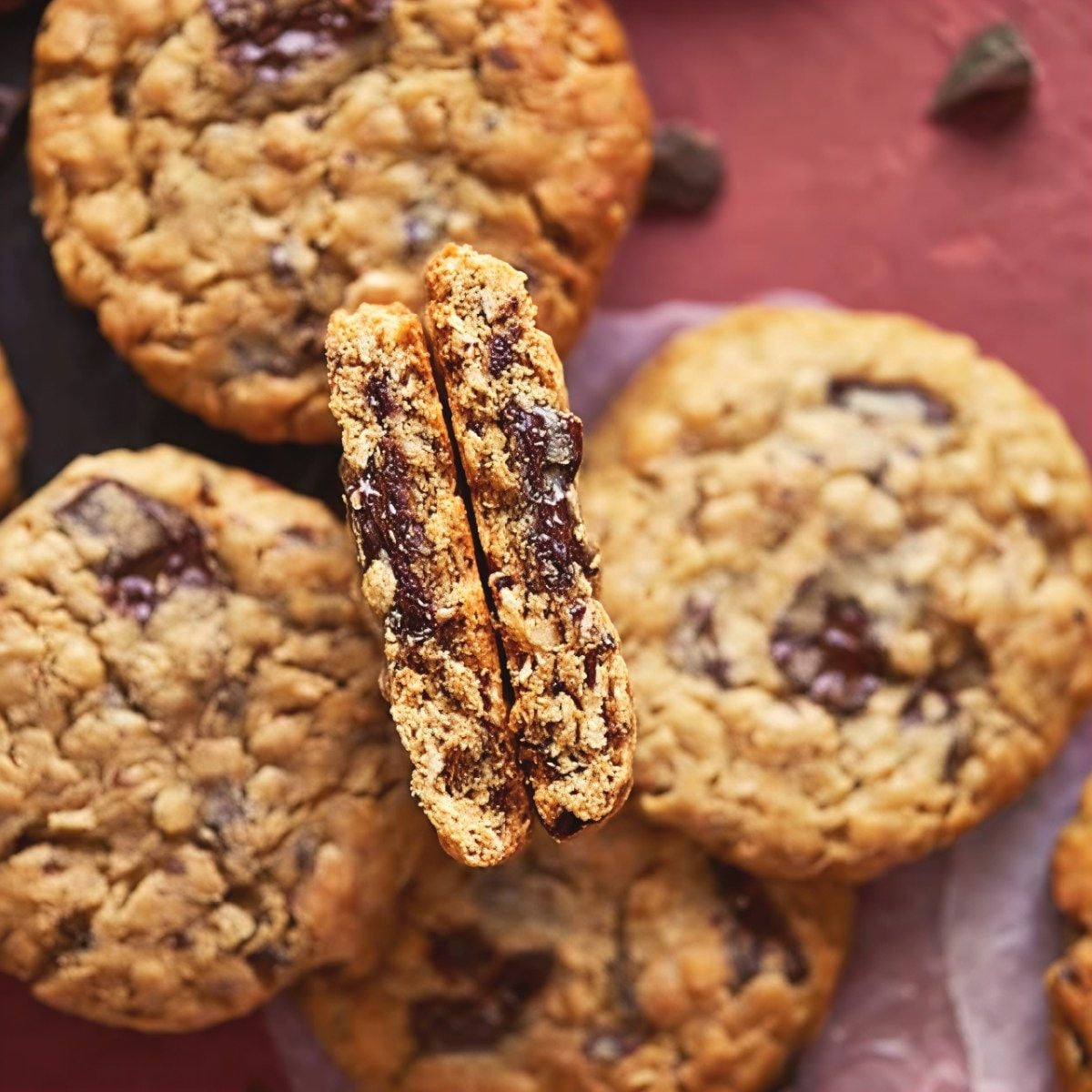 Two halves of a thick peanut butter oatmeal cookie held together to showcase a soft, chewy center packed with melted chocolate chips surrounded by round whole cookies on a surface.