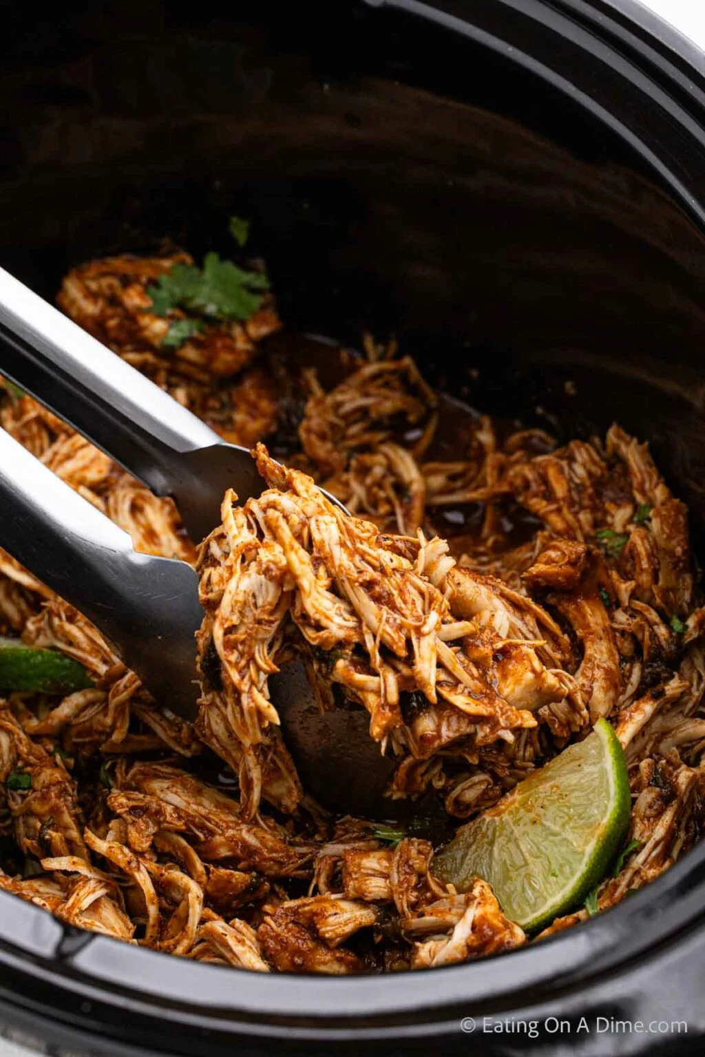 Tongs lifting seasoned shredded chicken with lime wedges out of a slow cooker, showcasing an effortless dump and go crockpot dinner (Lazy Dinner).