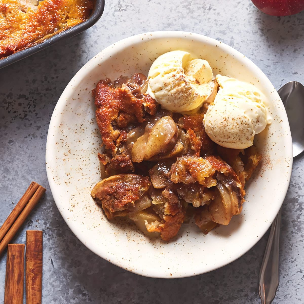 An overhead photograph of a serving of warm apple cobbler with a golden crust and spiced apples in a white bowl, topped with two scoops of vanilla ice cream and dusted with cinnamon.