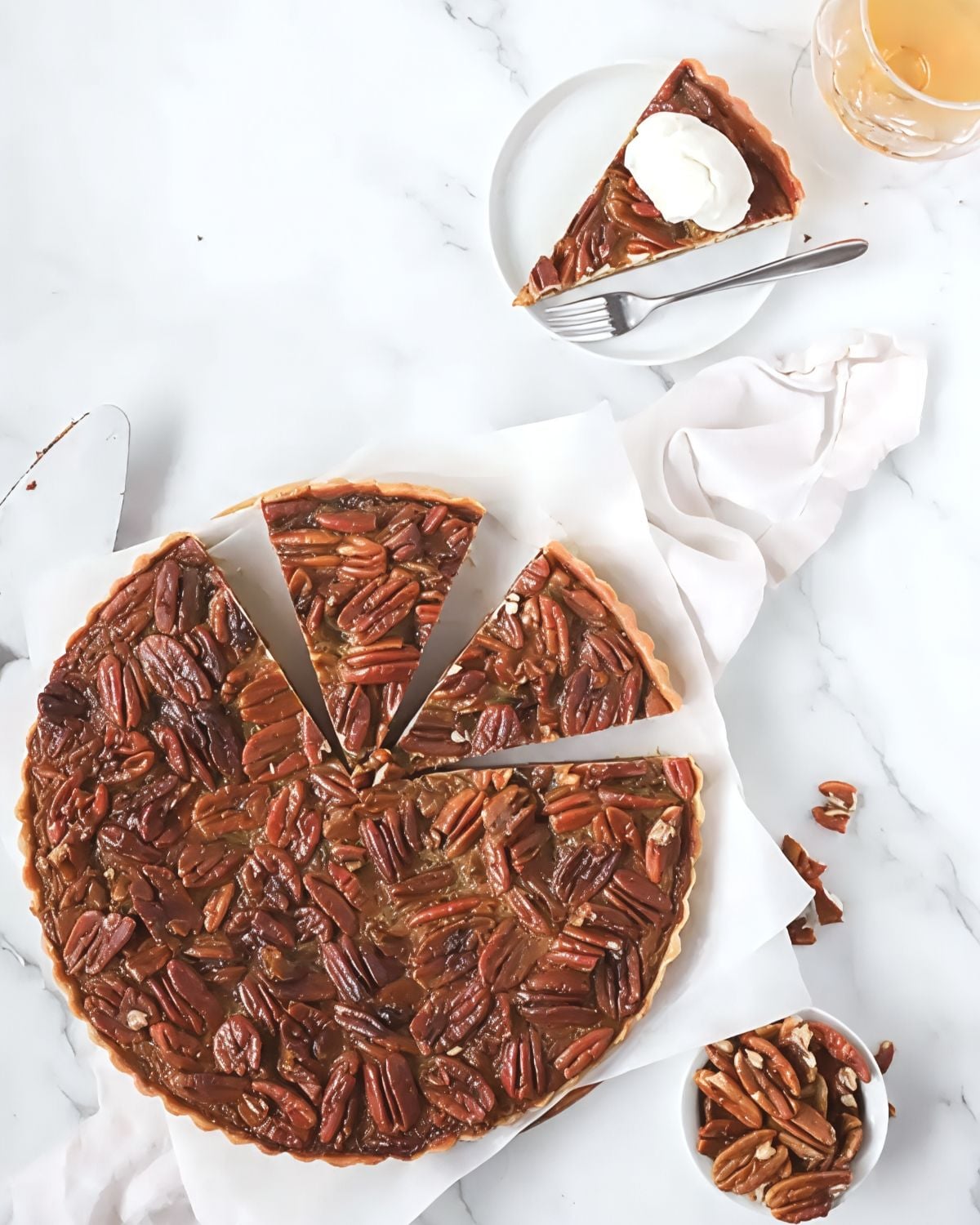 An overhead view of a sliced bourbon pecan tart with a fluted crust, showcasing a rich filling packed with toasted pecan halves alongside a plated slice topped with whipped cream.