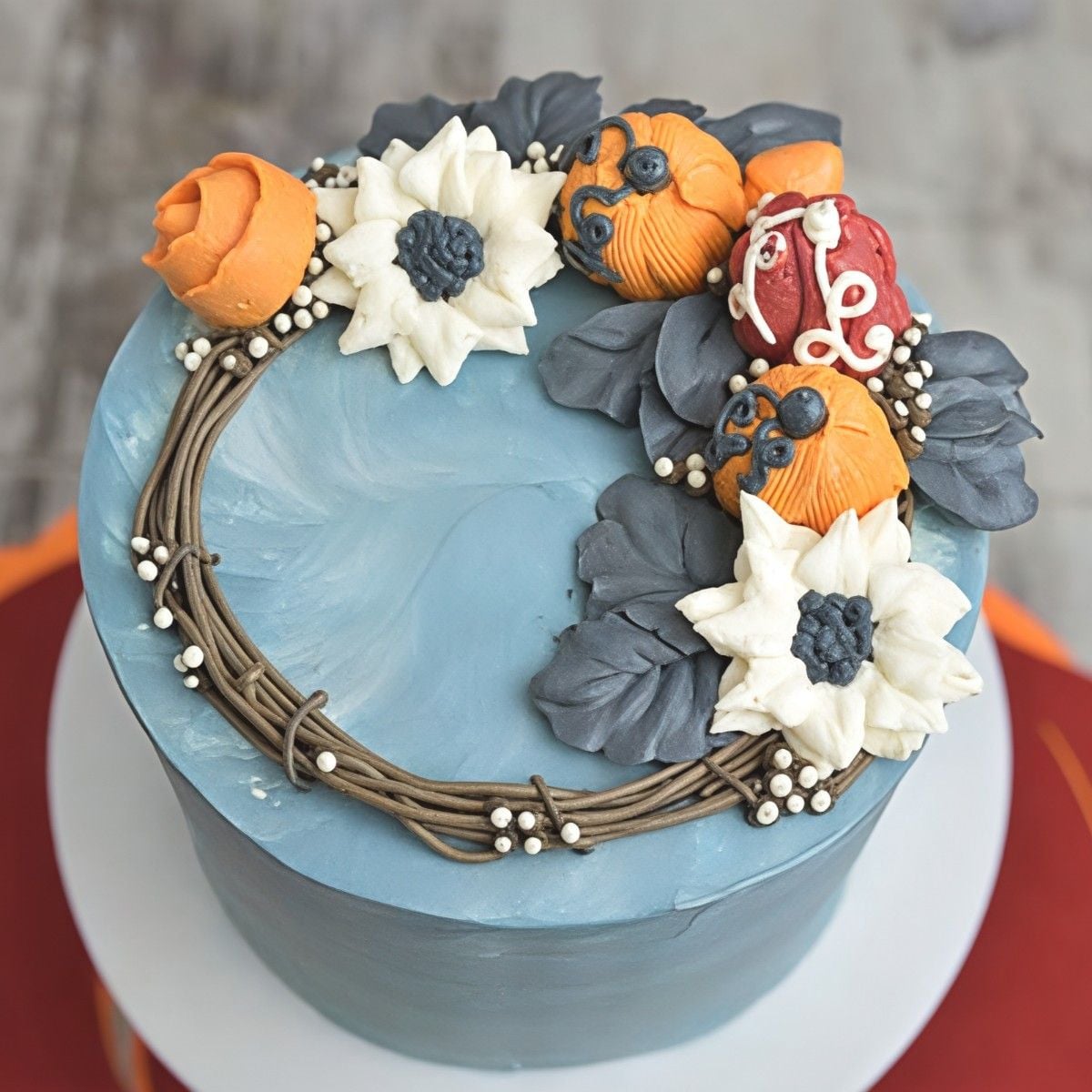 Smooth grey-blue frosted Thanksgiving cake with a slice removed to reveal yellow layers, topped with an intricate buttercream fall wreath featuring orange pumpkins and white flowers.