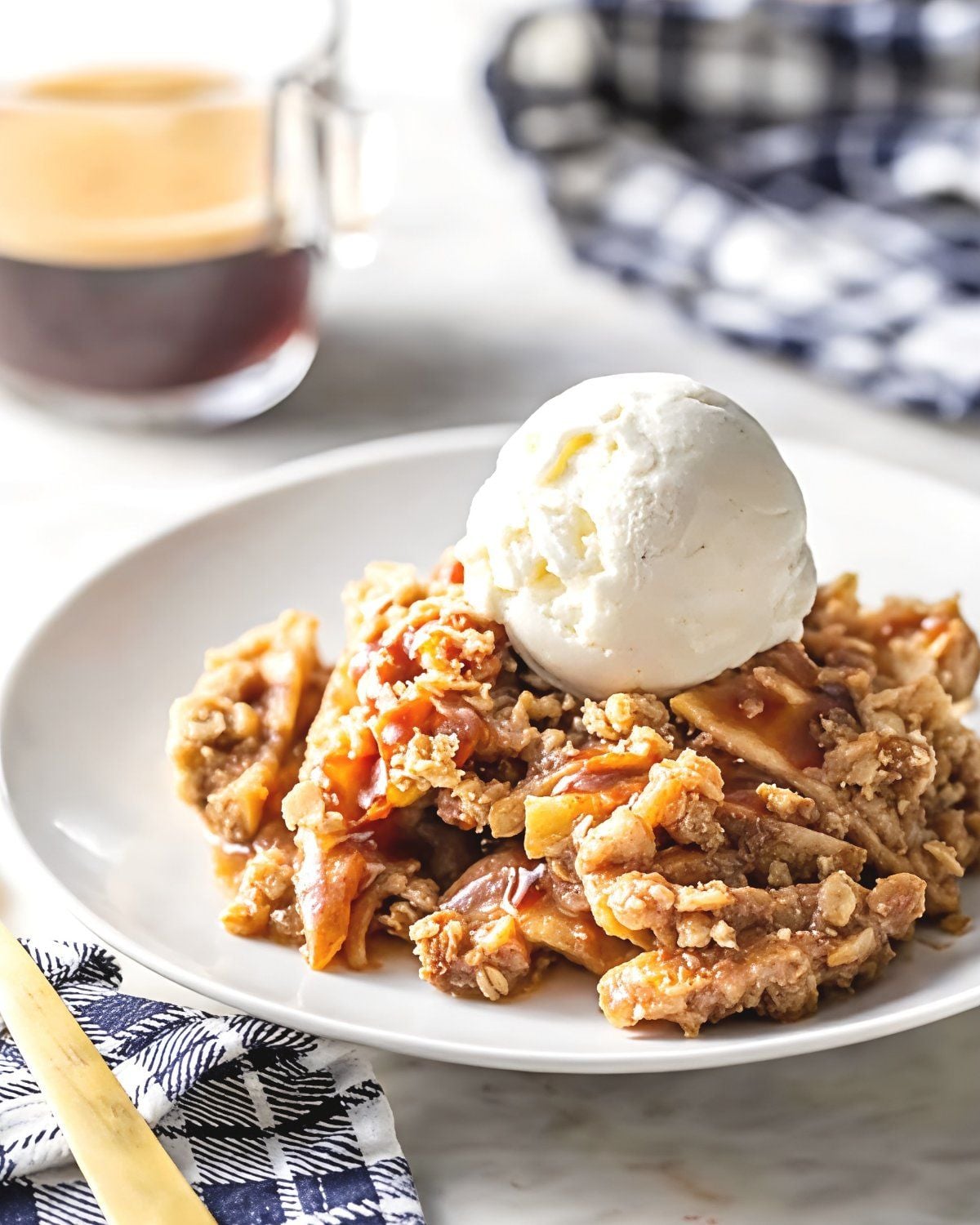 Warm caramel apple crisp layered with sliced cinnamon apples and a crunchy oat streusel topping, crowned with a scoop of vanilla ice cream on a white plate.