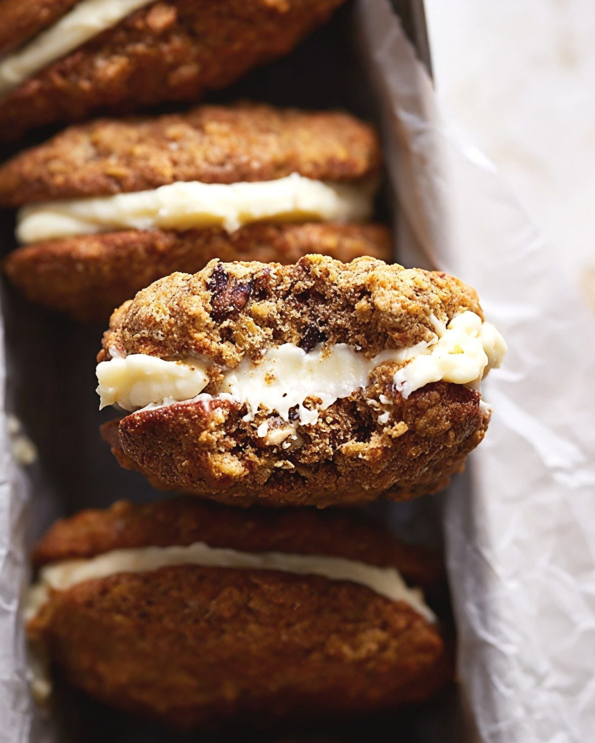 A row of carrot cake cookie sandwiches filled with rich cream cheese frosting packed in a paper-lined box, with a close-up on one sandwich revealing a soft, spiced cookie texture.