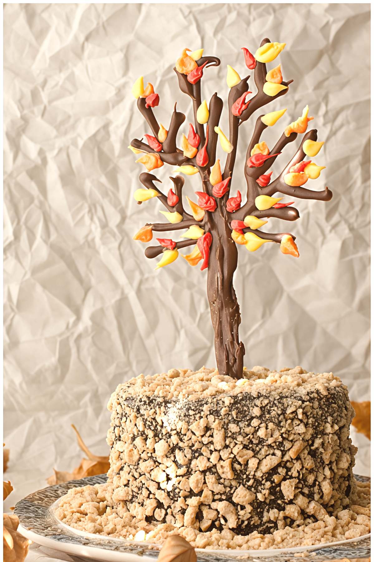 Rustic Thanksgiving cake coated in crushed cookie crumbs and topped with an edible chocolate tree decorated with colorful autumn leaf piping.