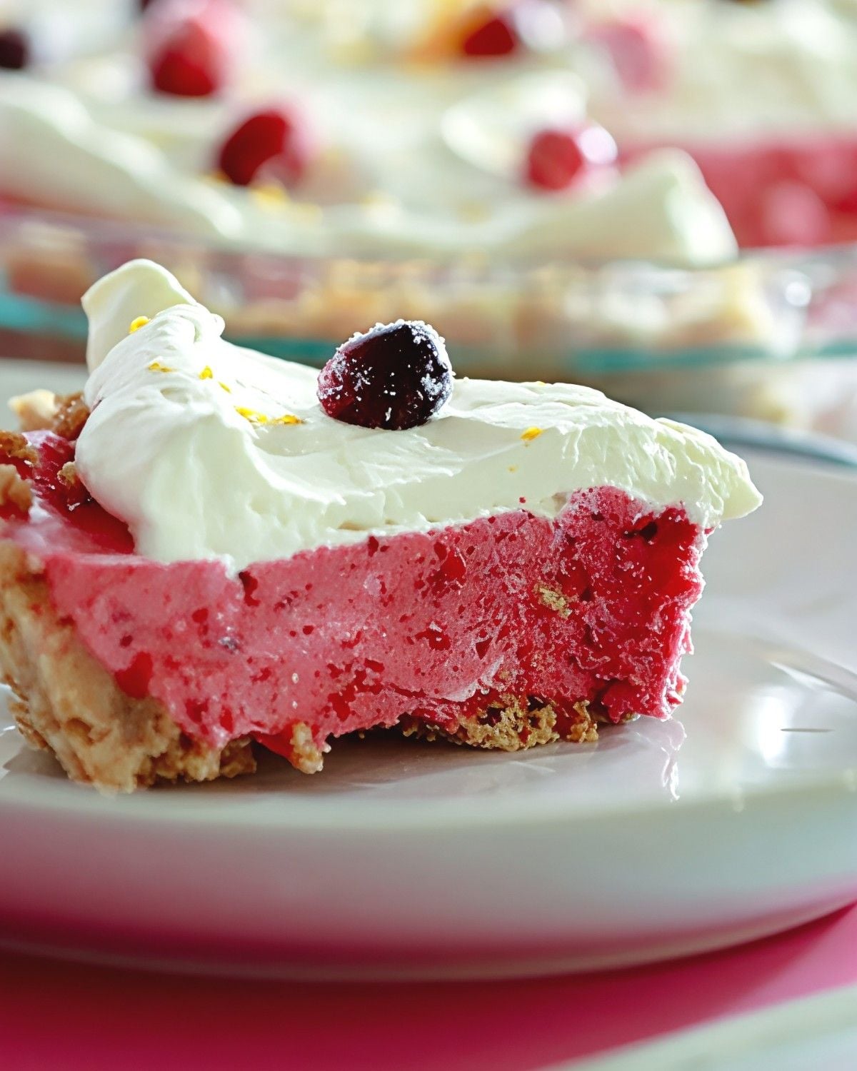A slice of airy cranberry chiffon pie on a graham cracker crust, topped with fluffy whipped cream, orange zest, and a sugared cranberry