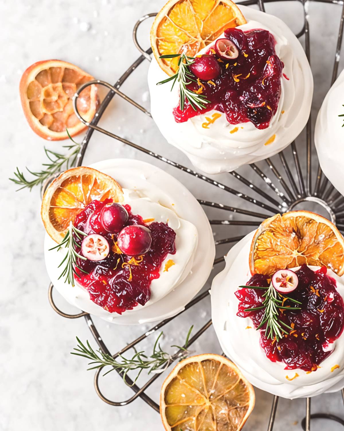 Crisp white meringue nests filled with whipped cream and topped with glossy tart cranberry compote, fresh rosemary sprigs, and dried orange slices on a wire cooling rack