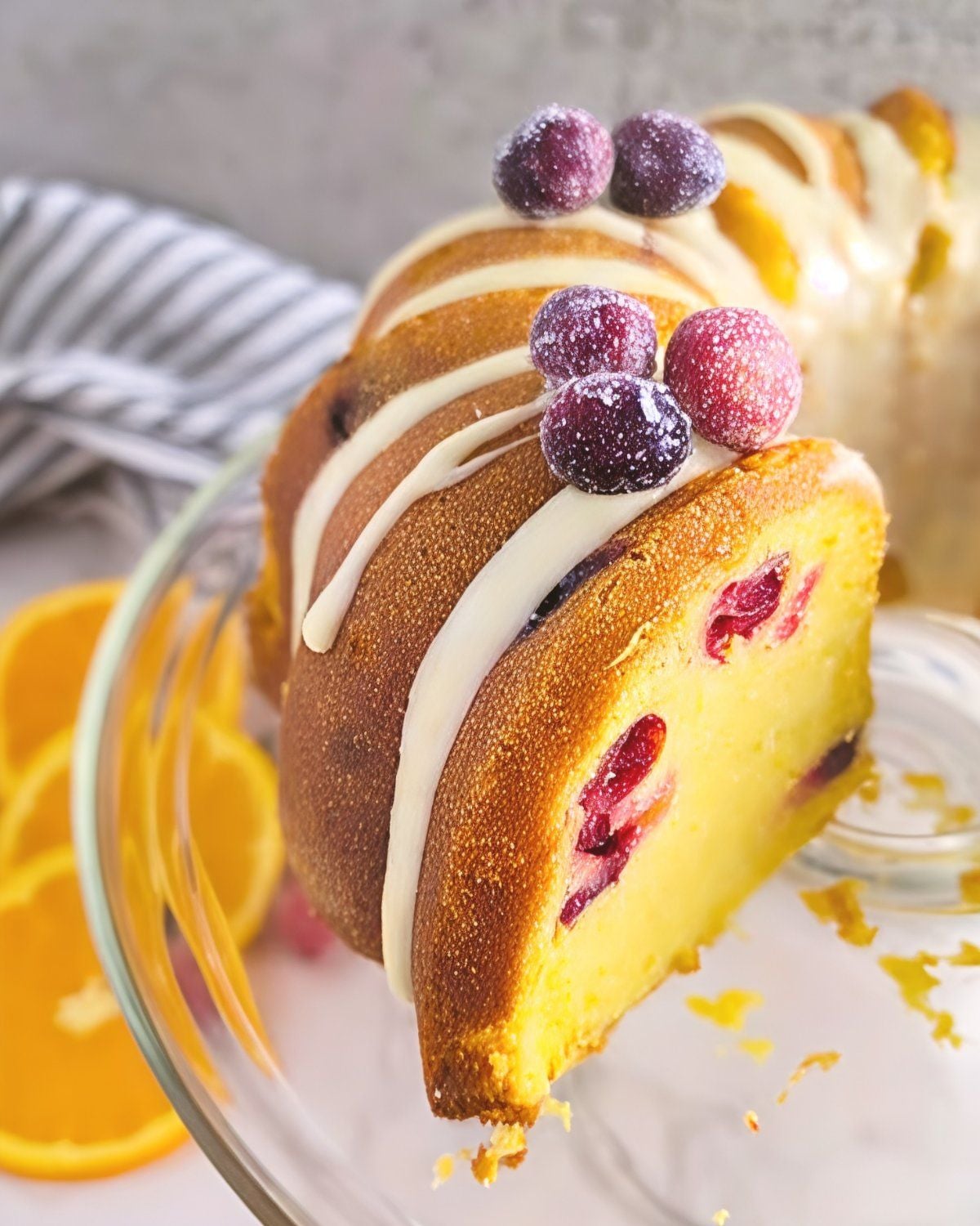 A slice of moist cranberry orange bundt cake studed with tart fresh cranberries, drizzled with sweet white glaze, and garnished with sugared cranberries on a glass cake stand