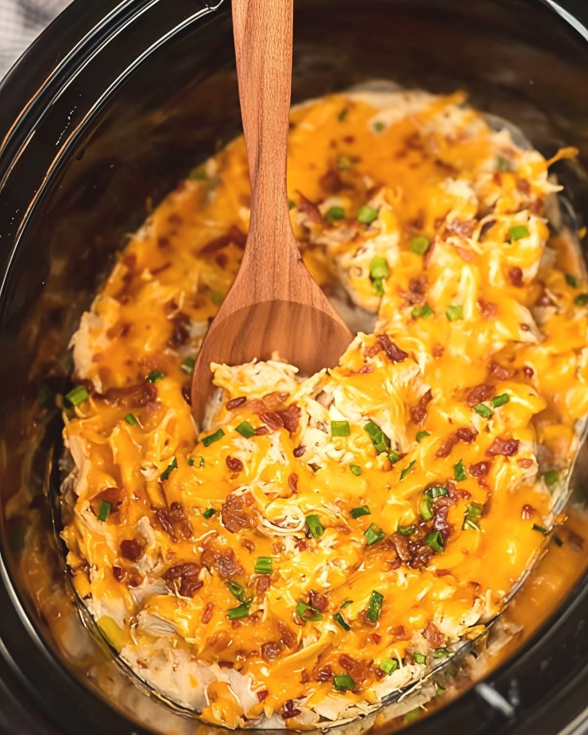 A wooden spoon scoops into cheesy crockpot crack chicken topped with bacon and green onions, showcasing a delicious dump and go crockpot dinners chicken recipe.