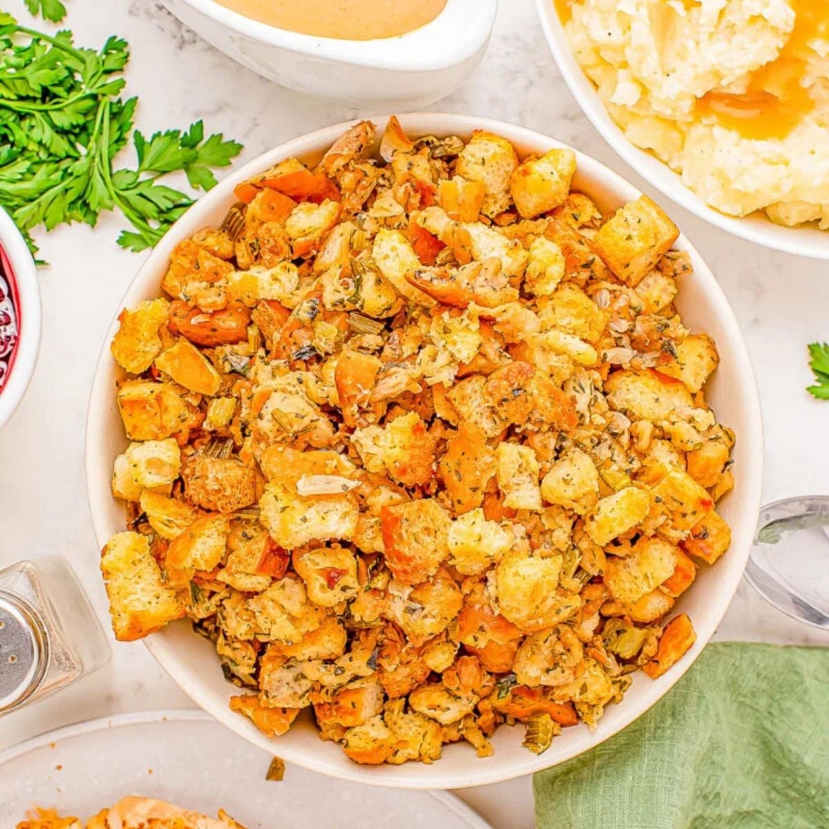 A bowl of golden crockpot stuffing thanksgiving recipe served alongside sliced turkey, mashed potatoes, and gravy.