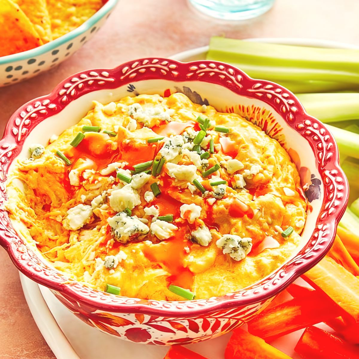 Creamy crockpot buffalo chicken dip topped with crumbled blue cheese and fresh chives in a decorative red bowl surrounded by fresh vegetables, perfect for crockpot thanksgiving recipes.