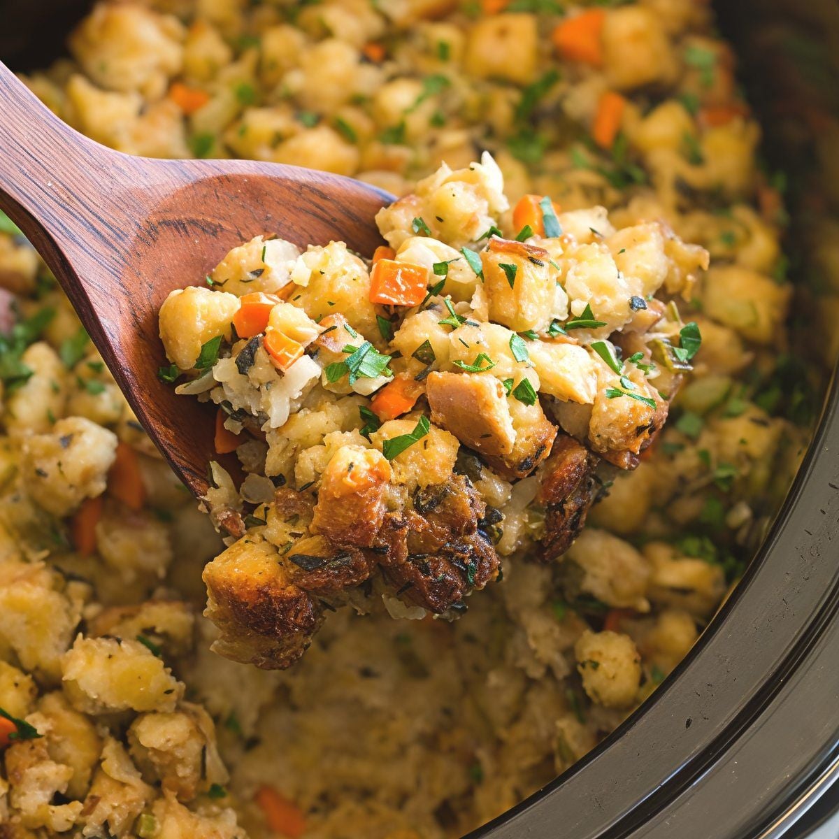 A wooden spoon scooping savory homemade stuffing out of a black slow cooker.