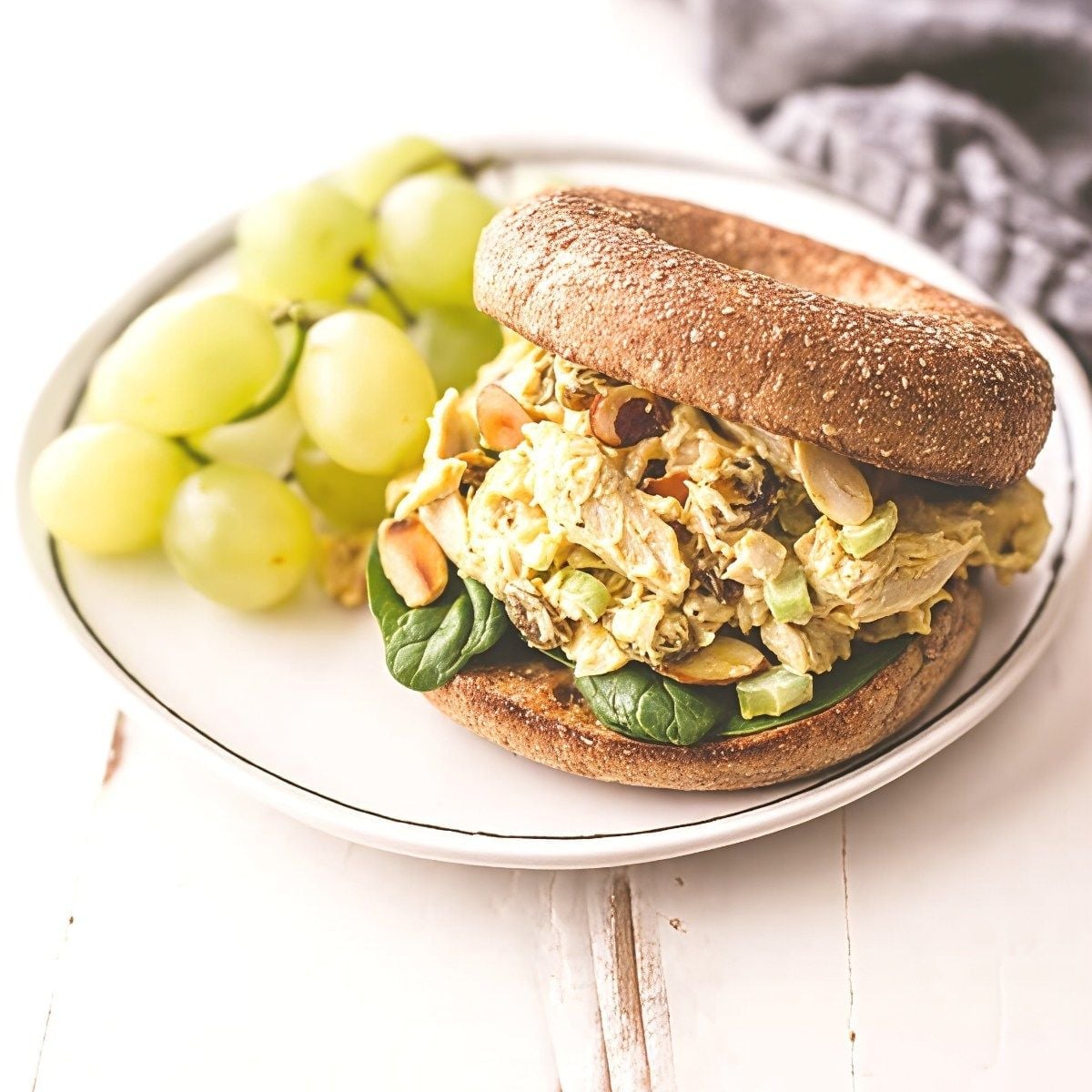 A whole wheat bagel sandwich loaded with creamy curried leftover turkey salad, sliced almonds, celery, and spinach, served on a plate with green grapes.