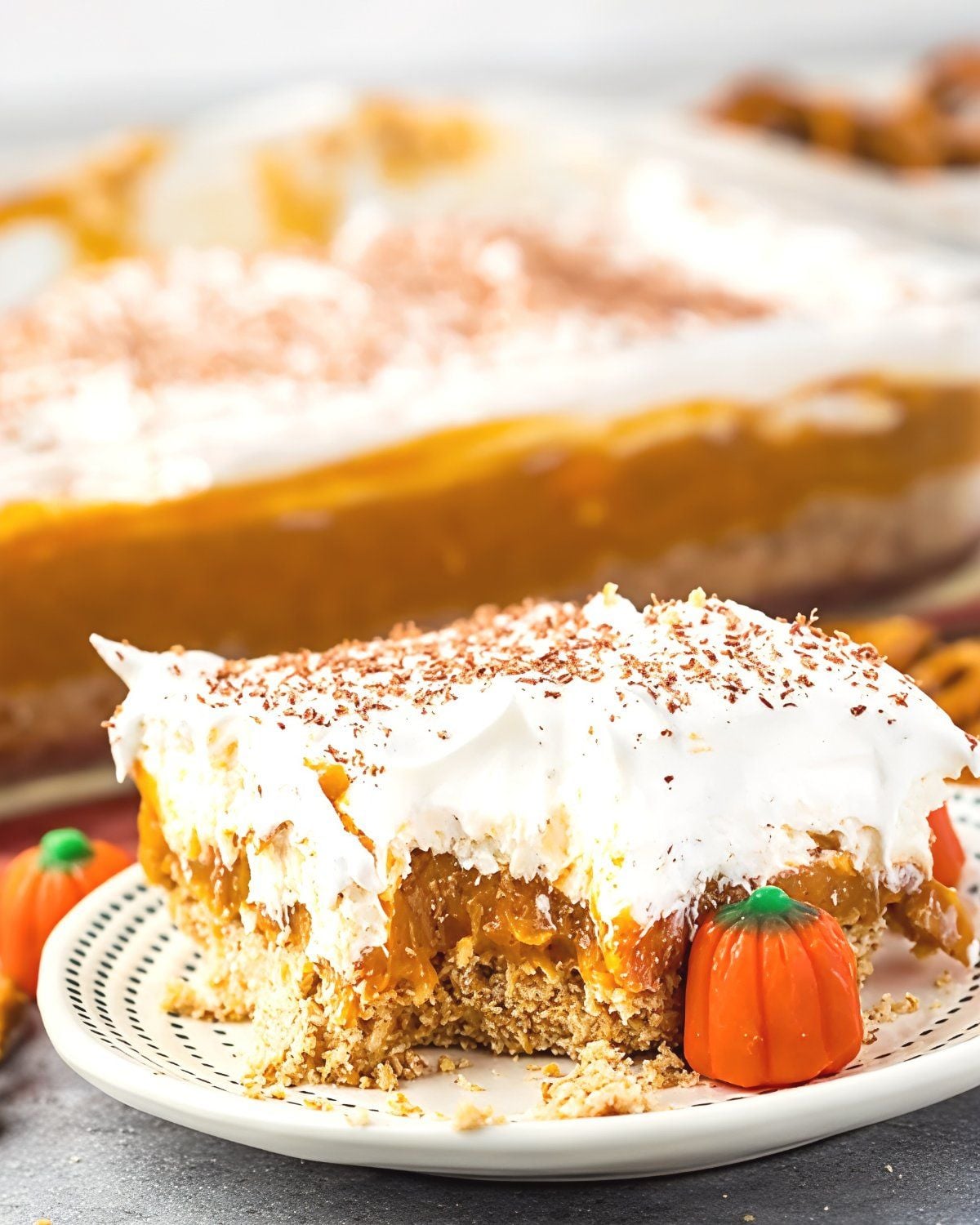 A slice of layered no-bake pumpkin dessert with a graham cracker crust, pumpkin filling, and whipped topping garnished with spice shavings and mini candy pumpkins for an easy Thanksgiving dessert.