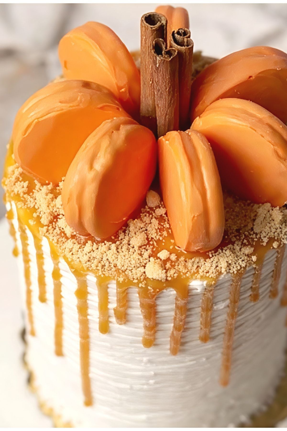 Elegant white frosted Thanksgiving cake with caramel drip, cookie crumbles, orange macarons arranged like a pumpkin, and whole cinnamon stick stems on top.
