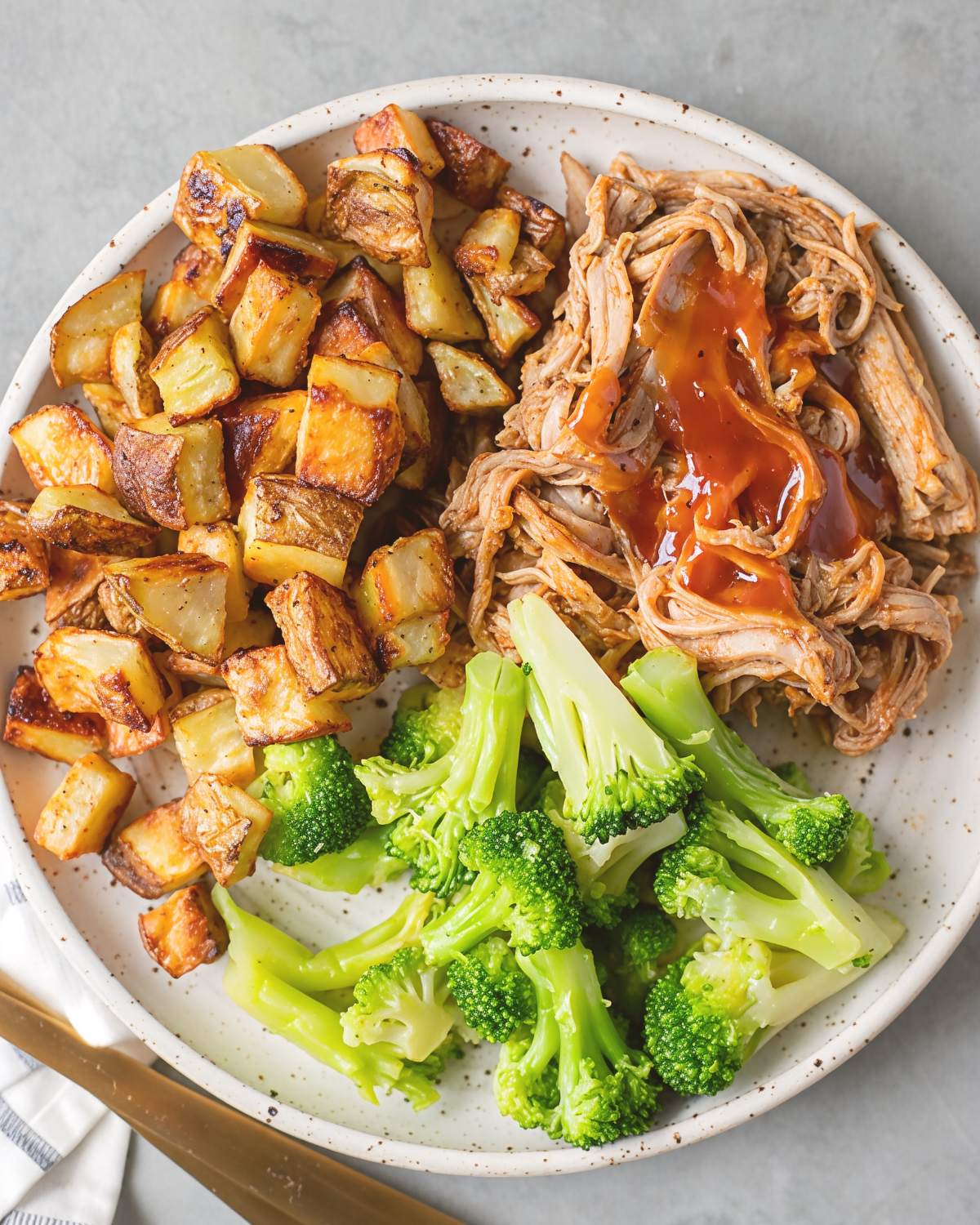 A healthy crockpot dinner plate featuring tender shredded pulled pork drizzled with savory barbecue sauce, crisp-tender steamed broccoli florets, and golden roasted potato cubes.