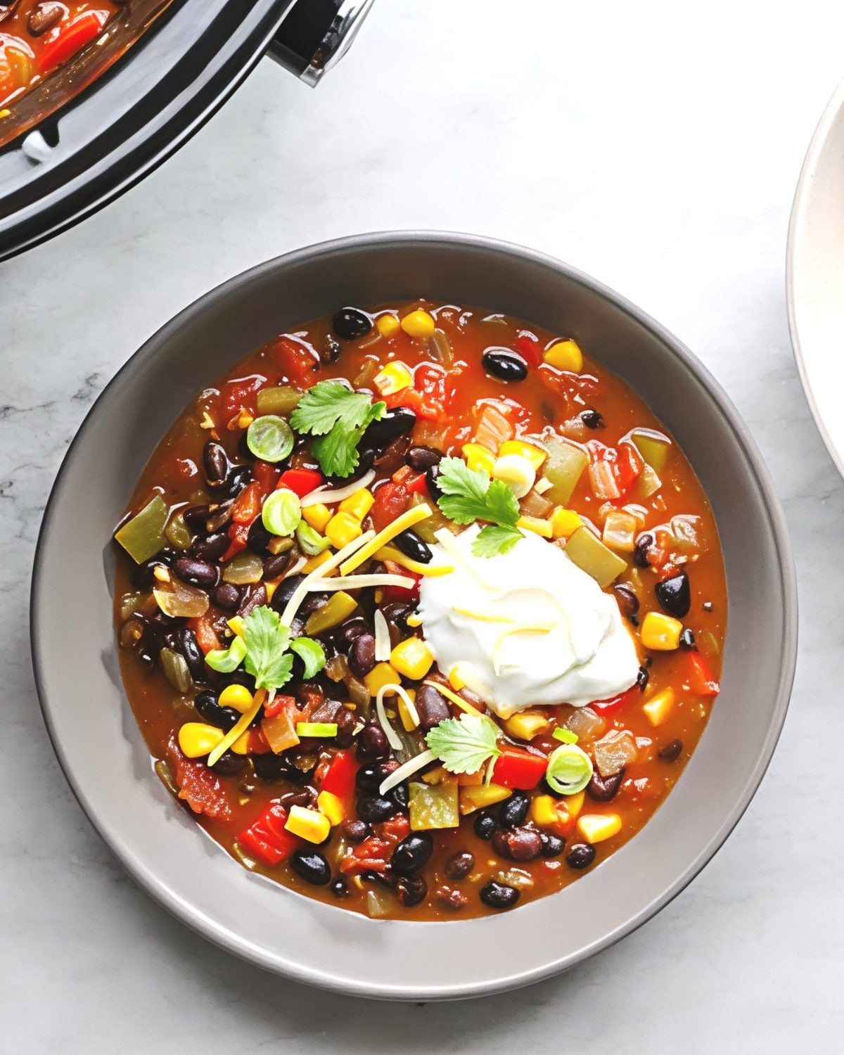 A bowl of vibrant crockpot black bean chili packed with sweet corn, diced red and green bell peppers, and tomatoes, topped with a dollop of sour cream, shredded cheese, green onions, and fresh cilantro leaves.