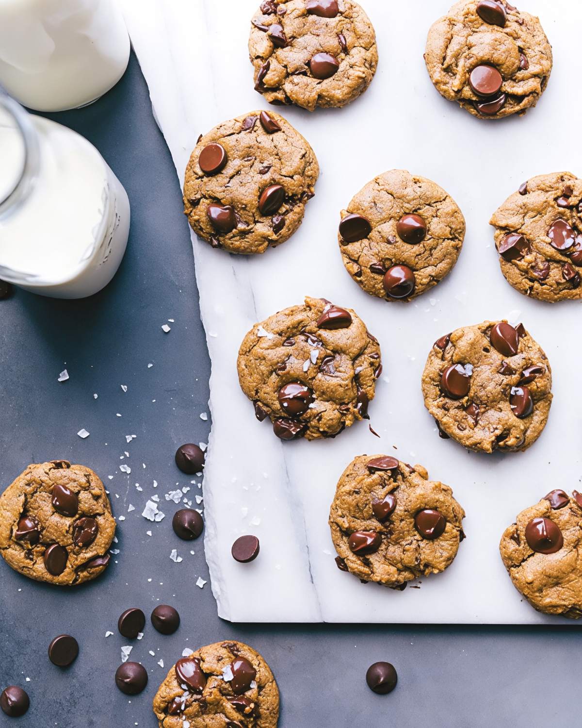 An overhead view of soft healthy pumpkin cookies studded with melted dark chocolate chips and sprinkled with flaky sea salt, arranged on marble parchment alongside bottles of milk.