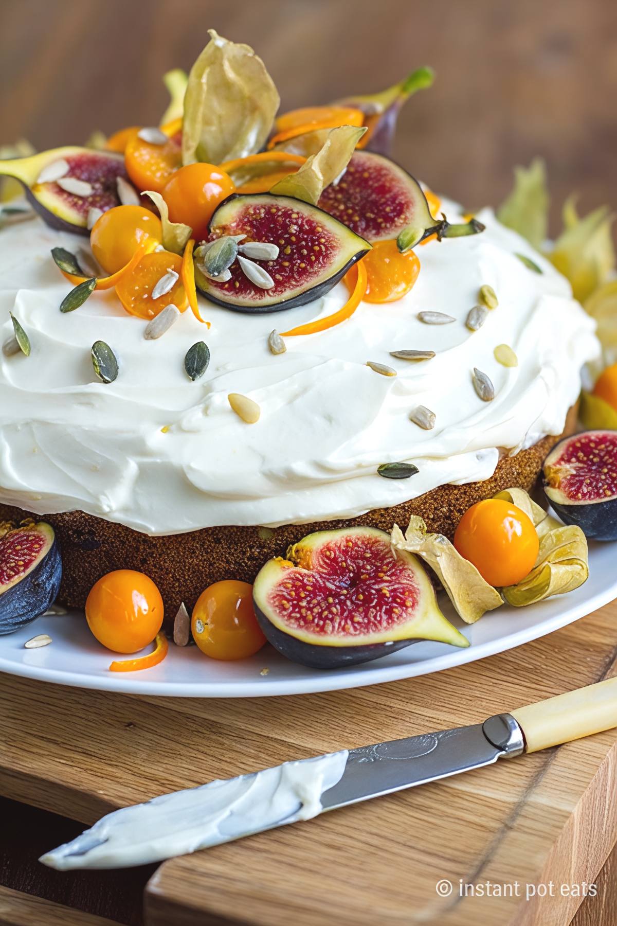 A round single-tier carrot cake topped with smooth white cream cheese frosting, sprinkled with pumpkin and sunflower seeds, and garnished with fresh sliced figs and orange cape gooseberries.