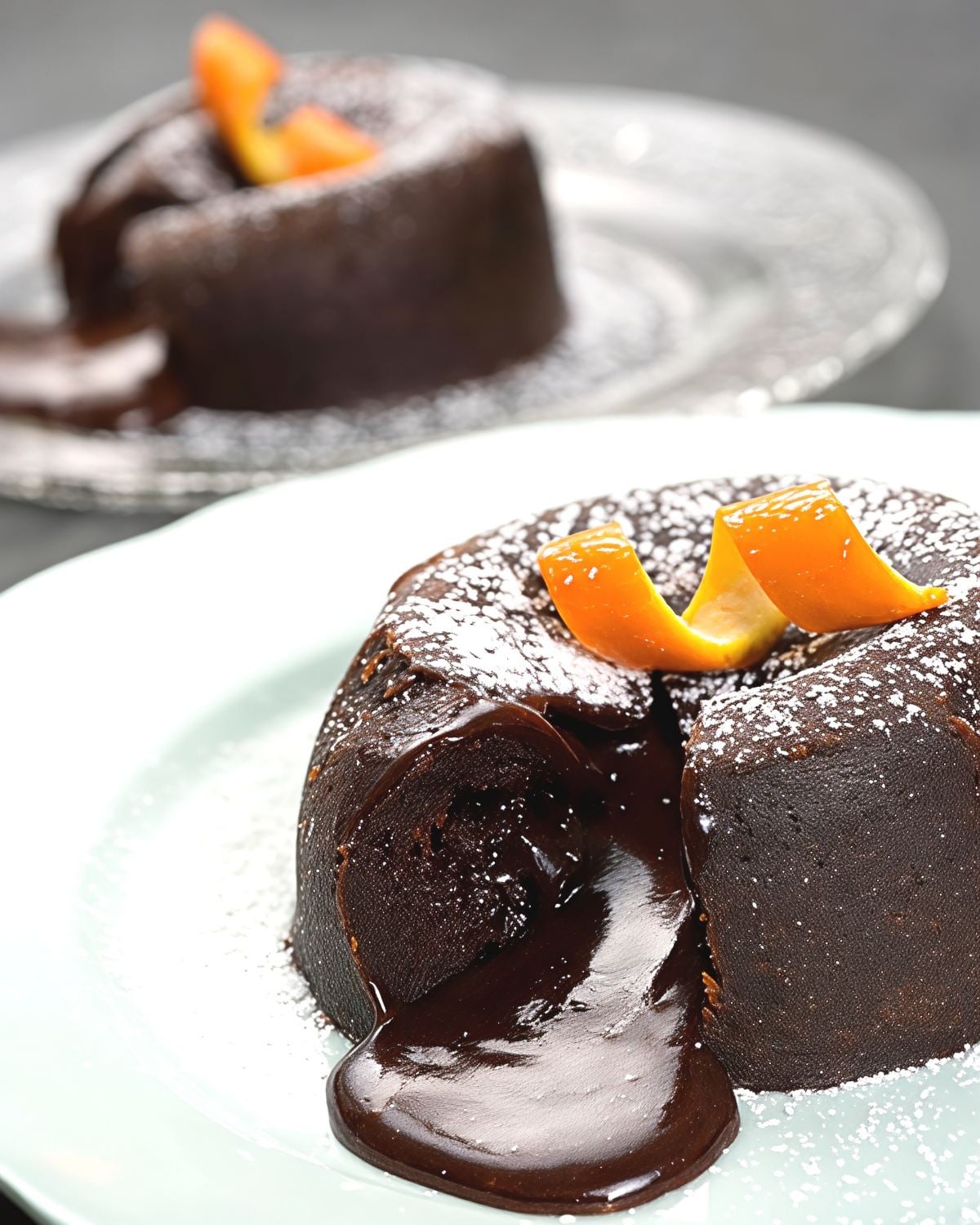 A decadent chocolate-orange lava cake on a plate dusted with powdered sugar, cut open to reveal warm chocolate fudge flowing out and garnished with a twist of orange peel.