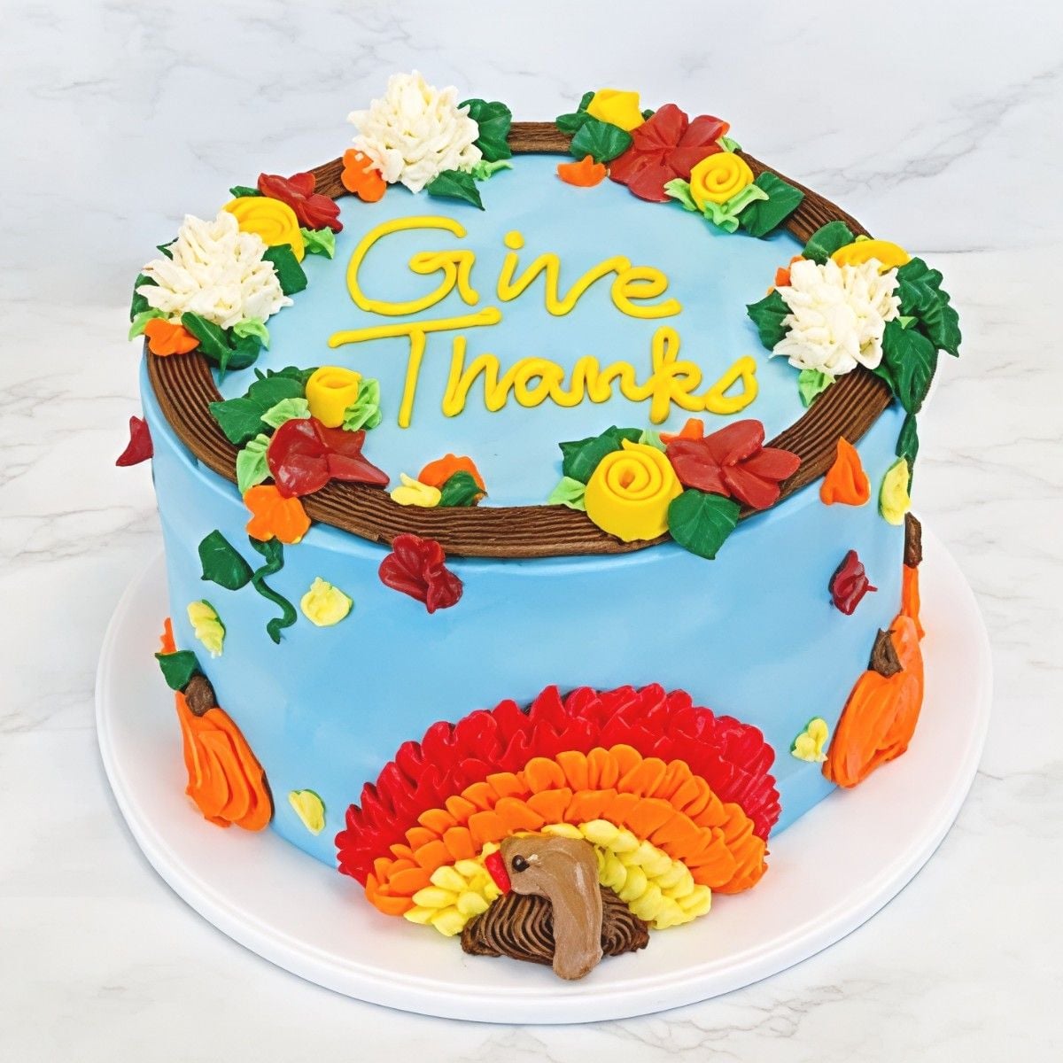 Light blue frosted Thanksgiving cake with yellow piped "Give Thanks" text, framed by a floral wreath border and a colorful buttercream turkey at the base.