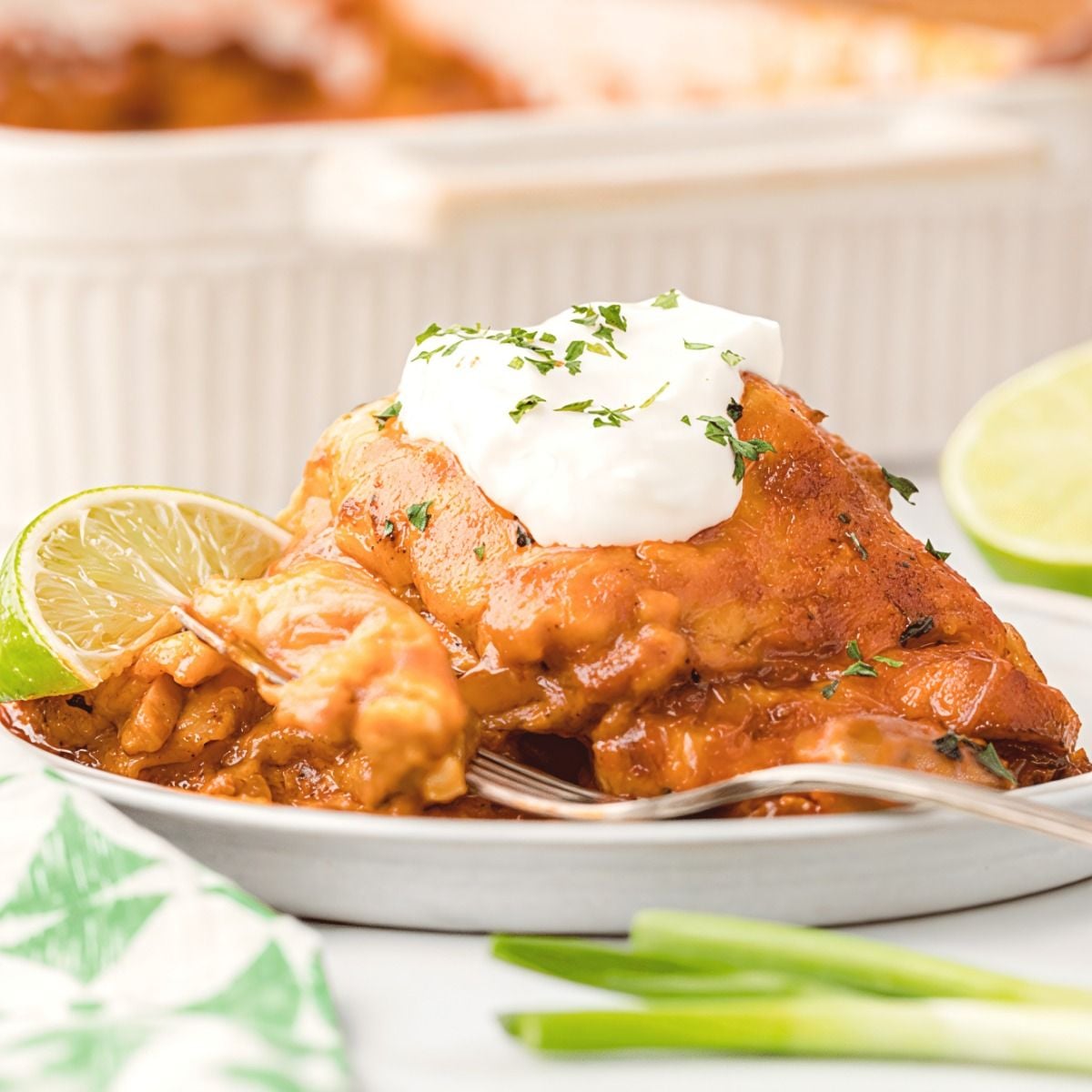 A serving of cheesy leftover turkey breast enchilada casserole topped with sour cream, cilantro, and fresh lime on a white plate.