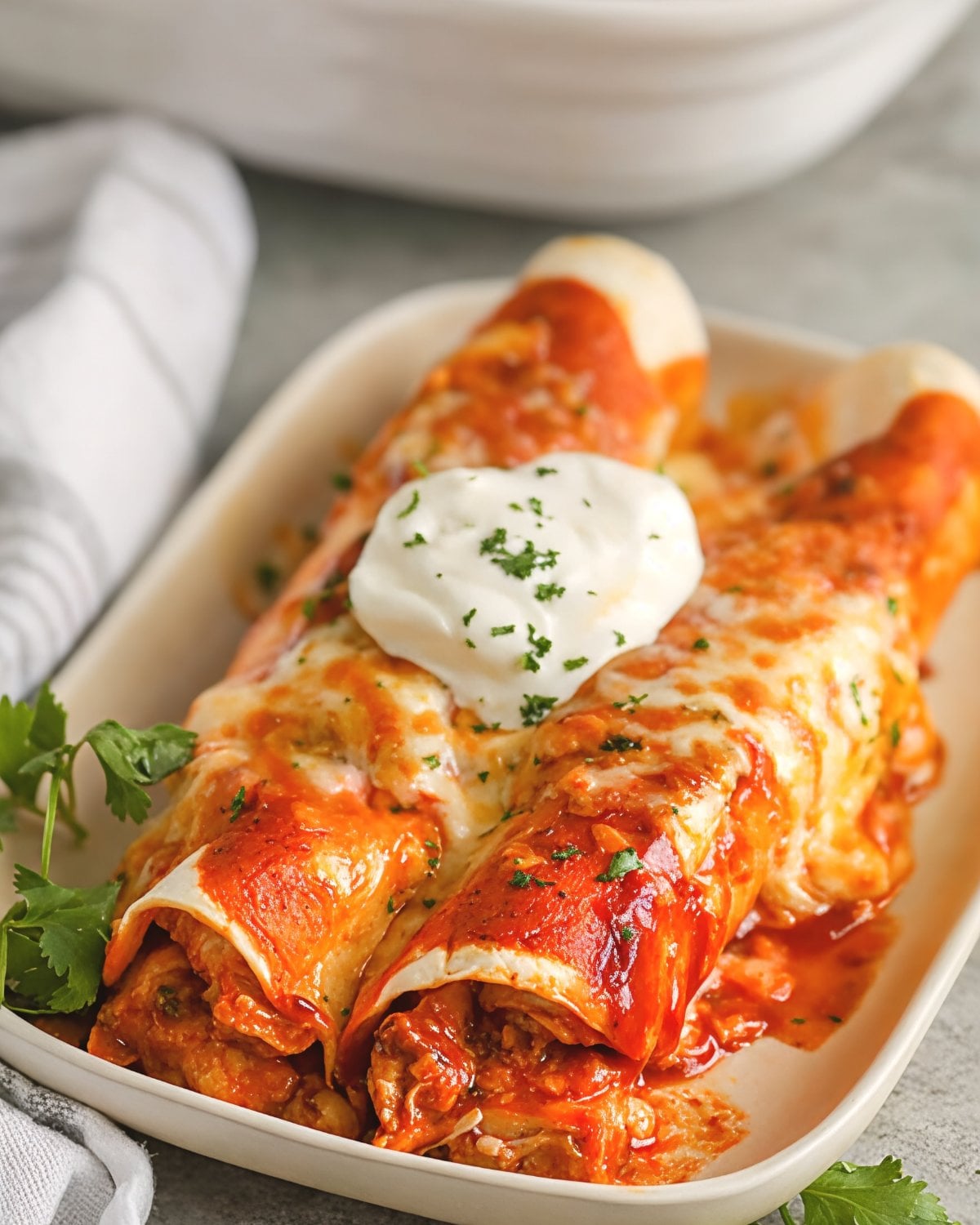 Cheesy leftover turkey enchiladas covered in red sauce and melted cheese, topped with sour cream and chopped cilantro on a plate.