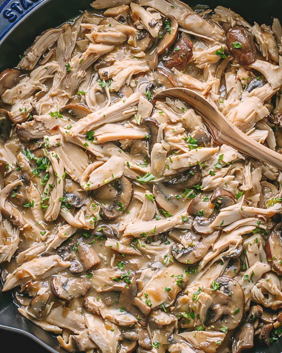 A wooden spoon rests in a pan filled with shredded leftover turkey simmered in rich gravy with sliced cremini mushrooms and fresh parsley.