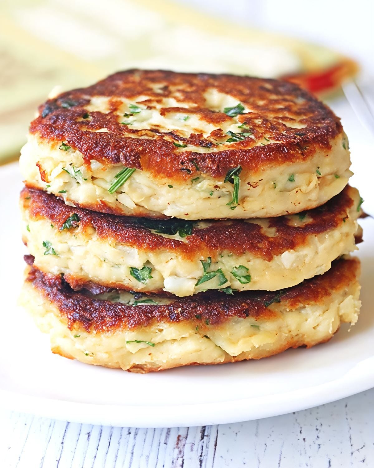A stack of three pan-fried golden brown leftover turkey patties flecked with fresh herbs served on a white plate.