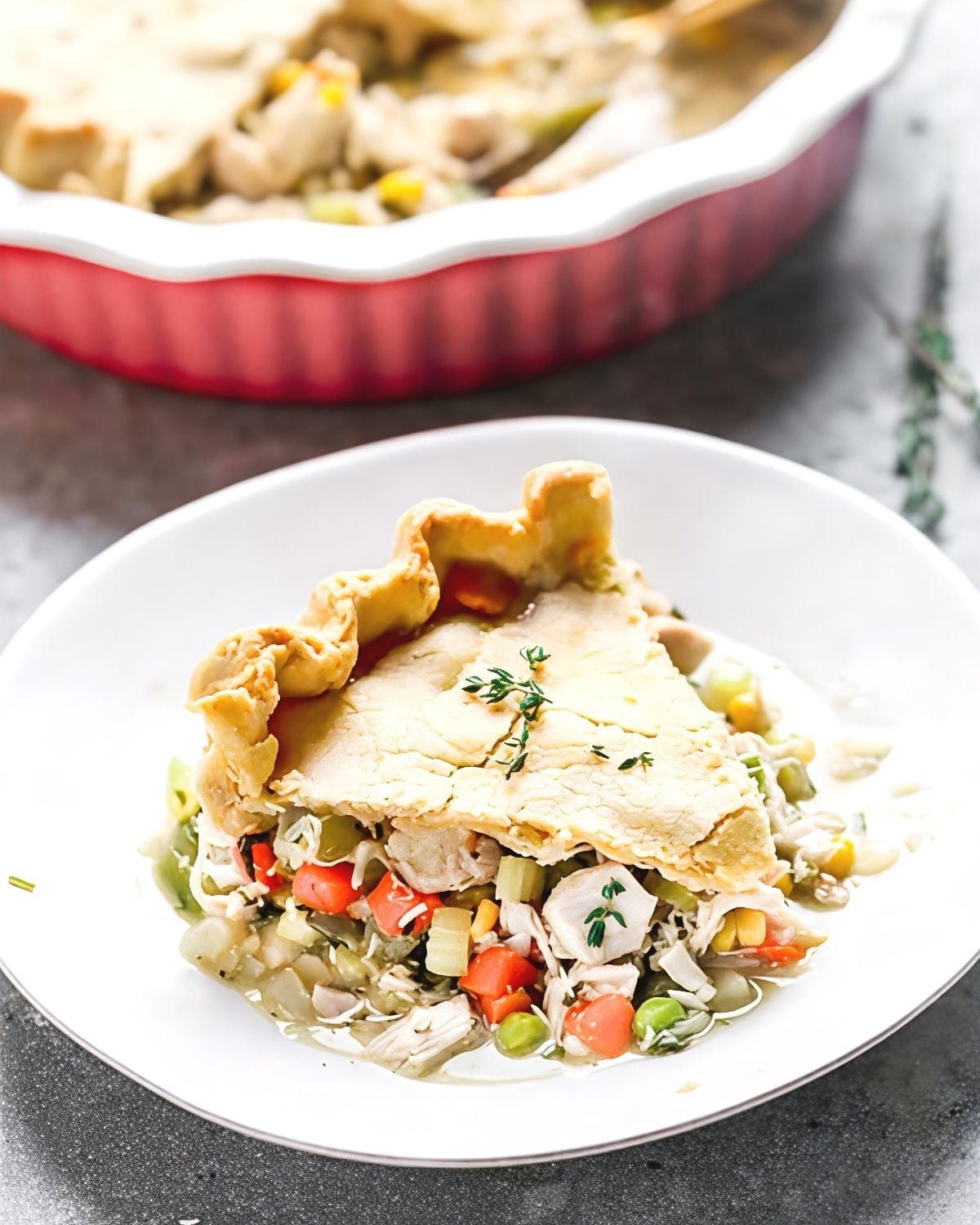 A golden slice of homemade turkey pot pie filled with shredded leftover turkey, carrots, peas, corn, and celery, topped with a flaky crimped crust and fresh thyme.
