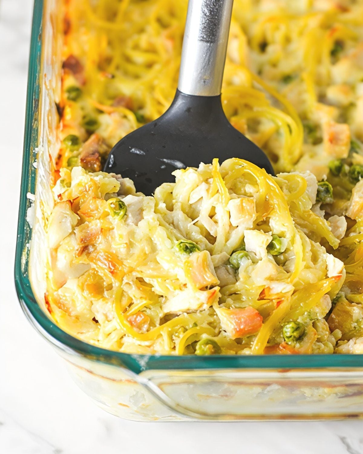 A black spatula scoops baked leftover turkey tetrazzini with spaghetti, diced turkey, green peas, and melted cheese from a glass casserole dish.