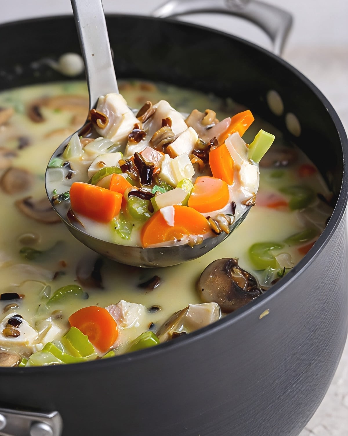Creamy leftover turkey wild rice soup with carrots, celery, and mushrooms scooped in a ladle over a cooking pot, perfect for cozy leftover turkey recipes.
