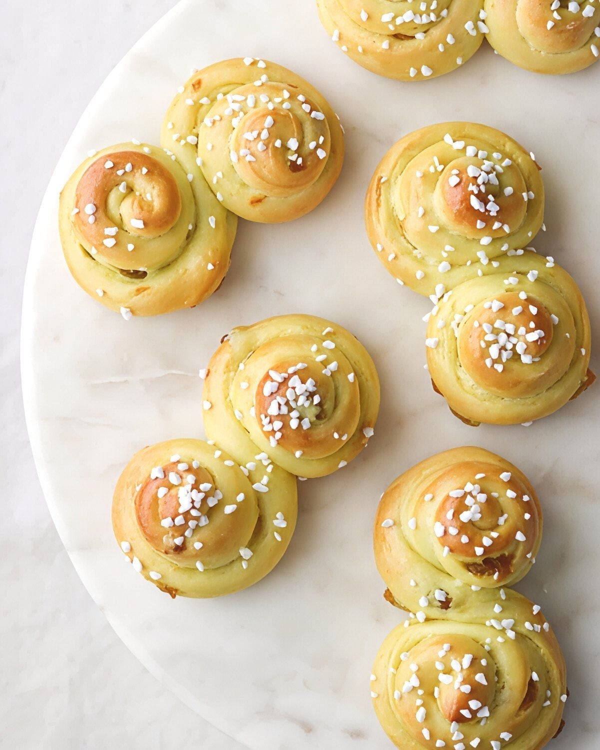 Golden Swedish Lussekatter saffron buns shaped in classic S-curves and sprinkled with pearl sugar, presented on a white marble platter as a European Christmas dessert.