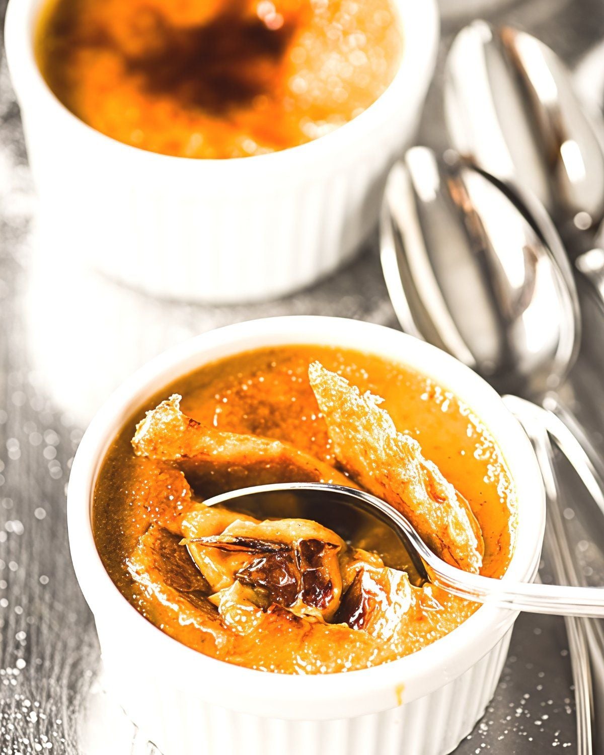 Silky maple bourbon pumpkin crème brûlée served in a white ramekin, featuring a golden caramelized sugar crust broken open with a silver dessert spoon
