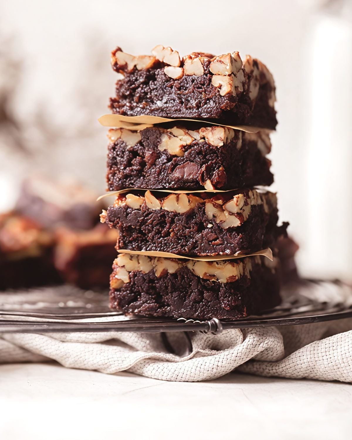A tall stack of fudgy chocolate pecan pie brownies layered with parchment paper, topped with a gooey, crunchy pecan layer and resting on a wire cooling rack