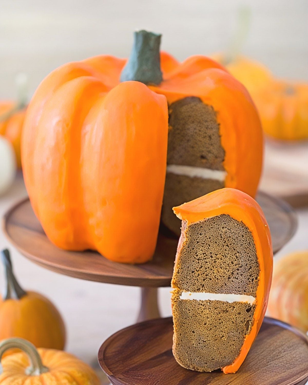 A festive double bundt pumpkin cake shaped like a real pumpkin with vibrant orange frosting, a fondant stem, and a white cream filling layer, with one slice served on a wooden plate