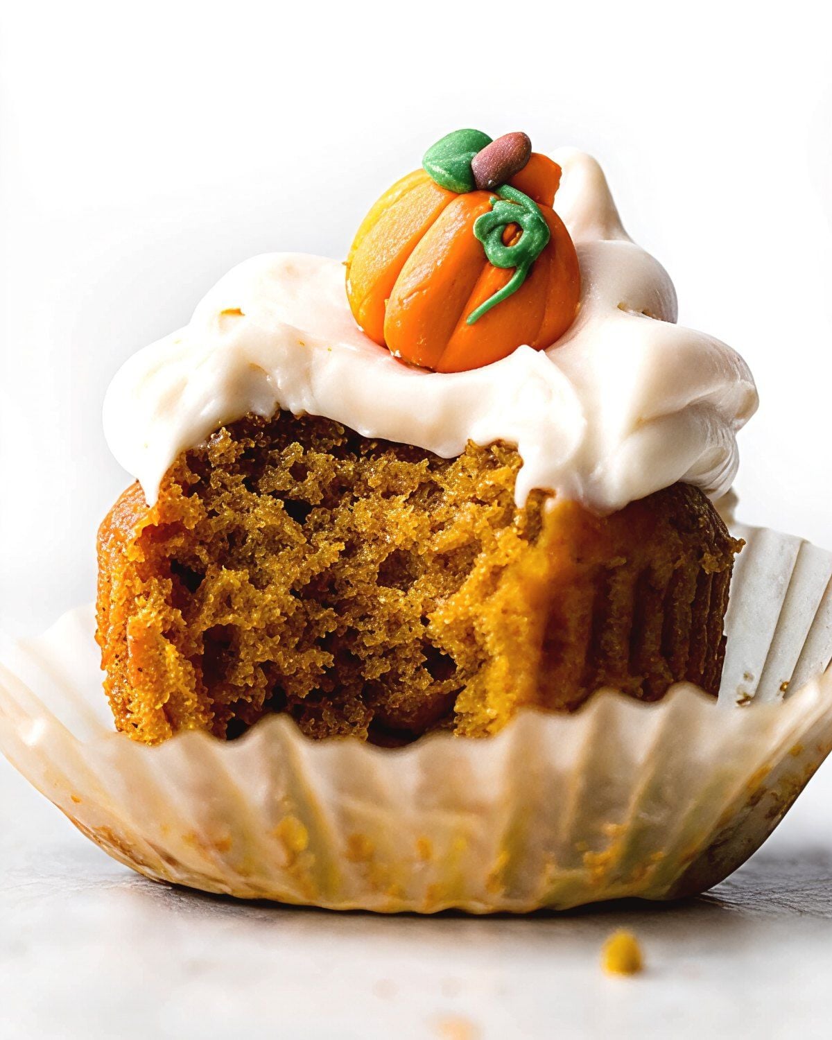 A moist spiced pumpkin cupcake with a bite taken out, unwrapped in a paper liner and topped with smooth cream cheese frosting and a cute mini orange pumpkin decoration