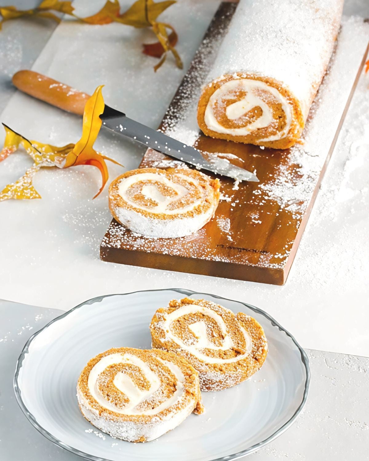 Sliced spiced pumpkin cake roll filled with rich cream cheese filling, lightly dusted with powdered sugar and arranged on a wooden board alongside autumn leaves.