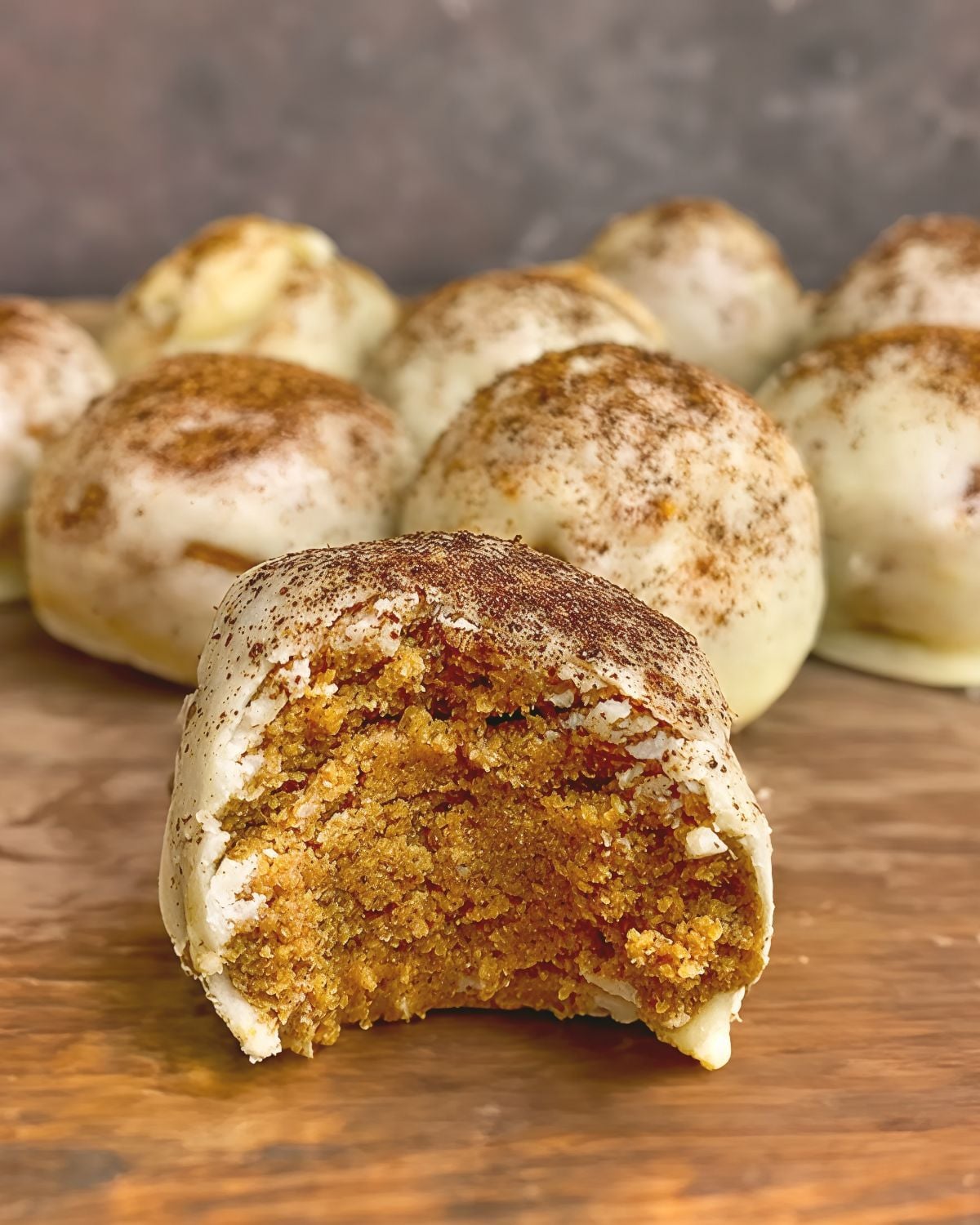 A close-up of no-bake pumpkin spice latte protein truffles coated in white chocolate and dusted with cinnamon, with a bite taken out of one to reveal its soft, spiced pumpkin filling.