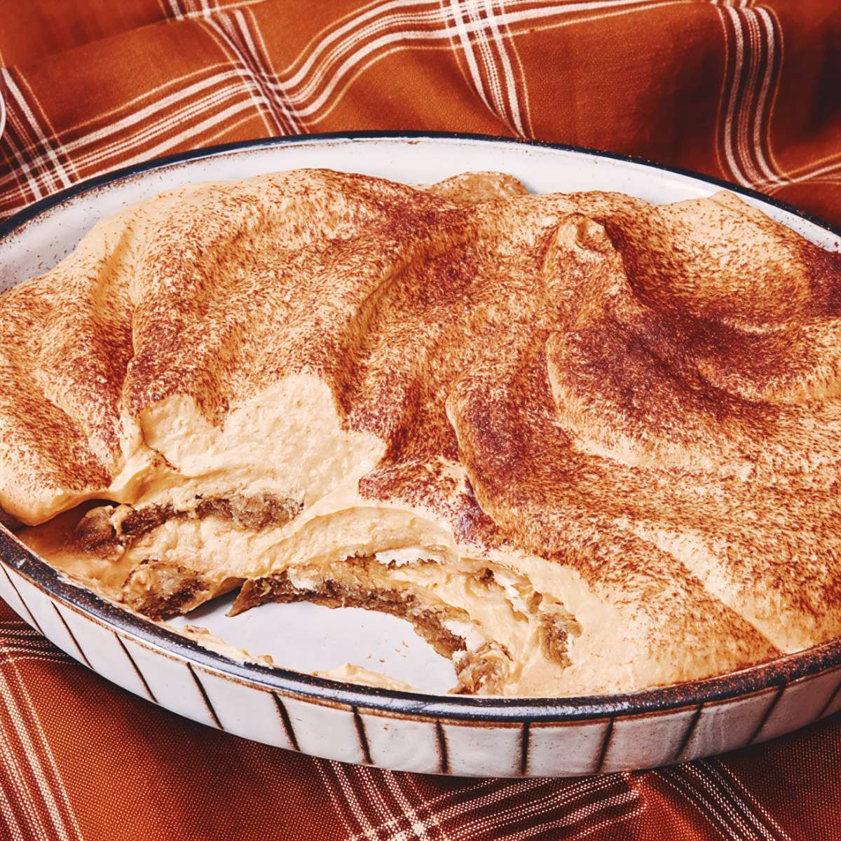 A rustic ceramic dish filled with creamy pumpkin tiramisu showcasing layers of espresso-soaked ladyfingers, spiced mascarpone cream, and a generous dusting of cocoa powder