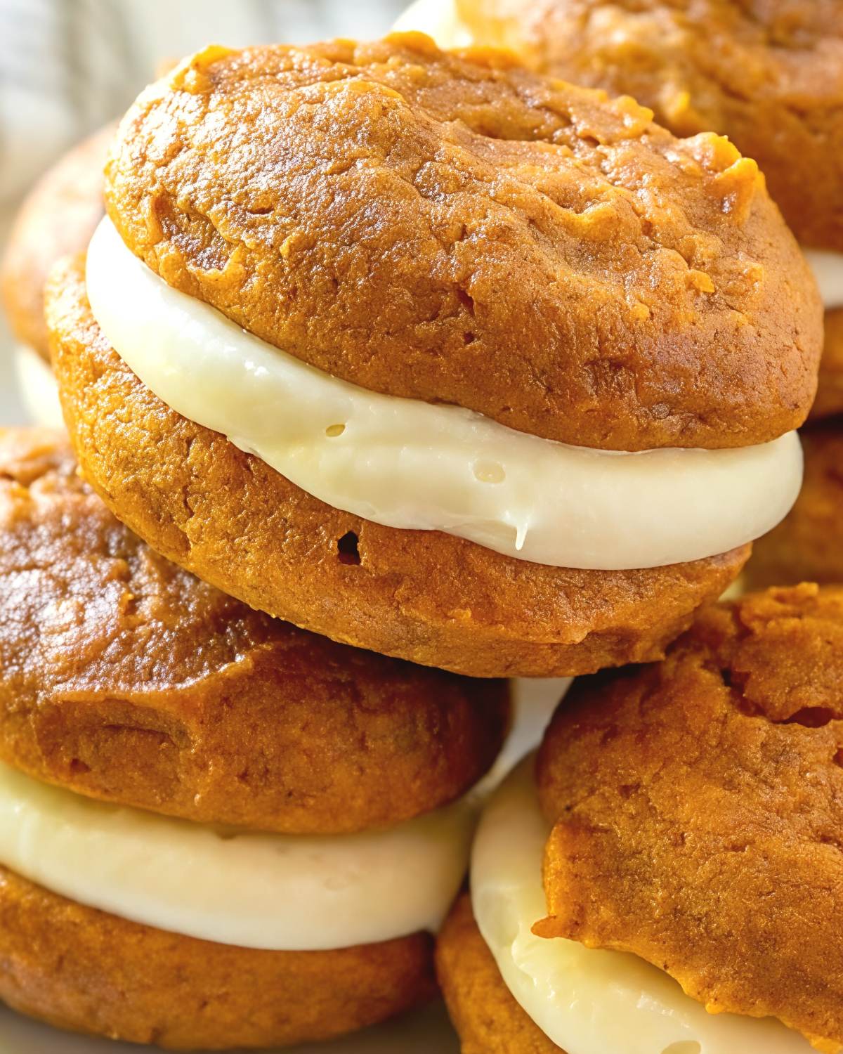 A close-up stack of soft, cake-like pumpkin whoopie pies generously filled with smooth, sweet cream cheese frosting