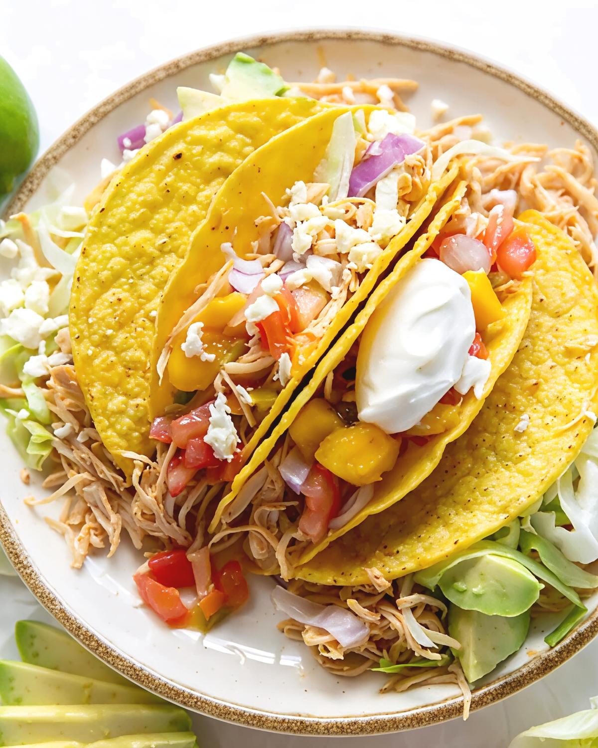Two crispy yellow corn tortillas on a white plate, filled with shredded crockpot chicken, fresh mango salsa, feta cheese, and sour cream, make for an easy and delicious dump and go dinner idea.