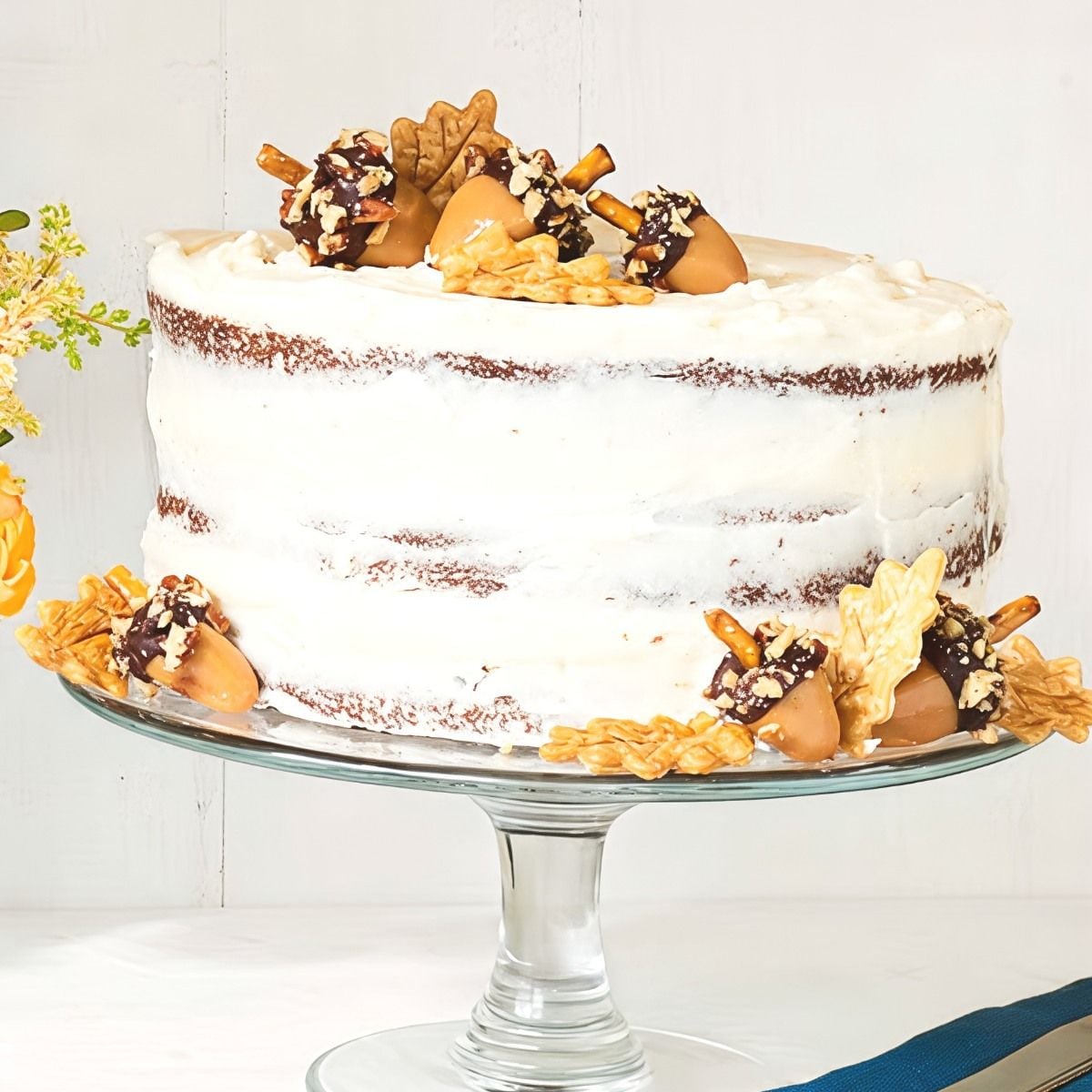Rustic Thanksgiving spice layer cake lightly coated with white frosting and decorated with edible chocolate-dipped pretzel acorns and pie crust autumn leaves.